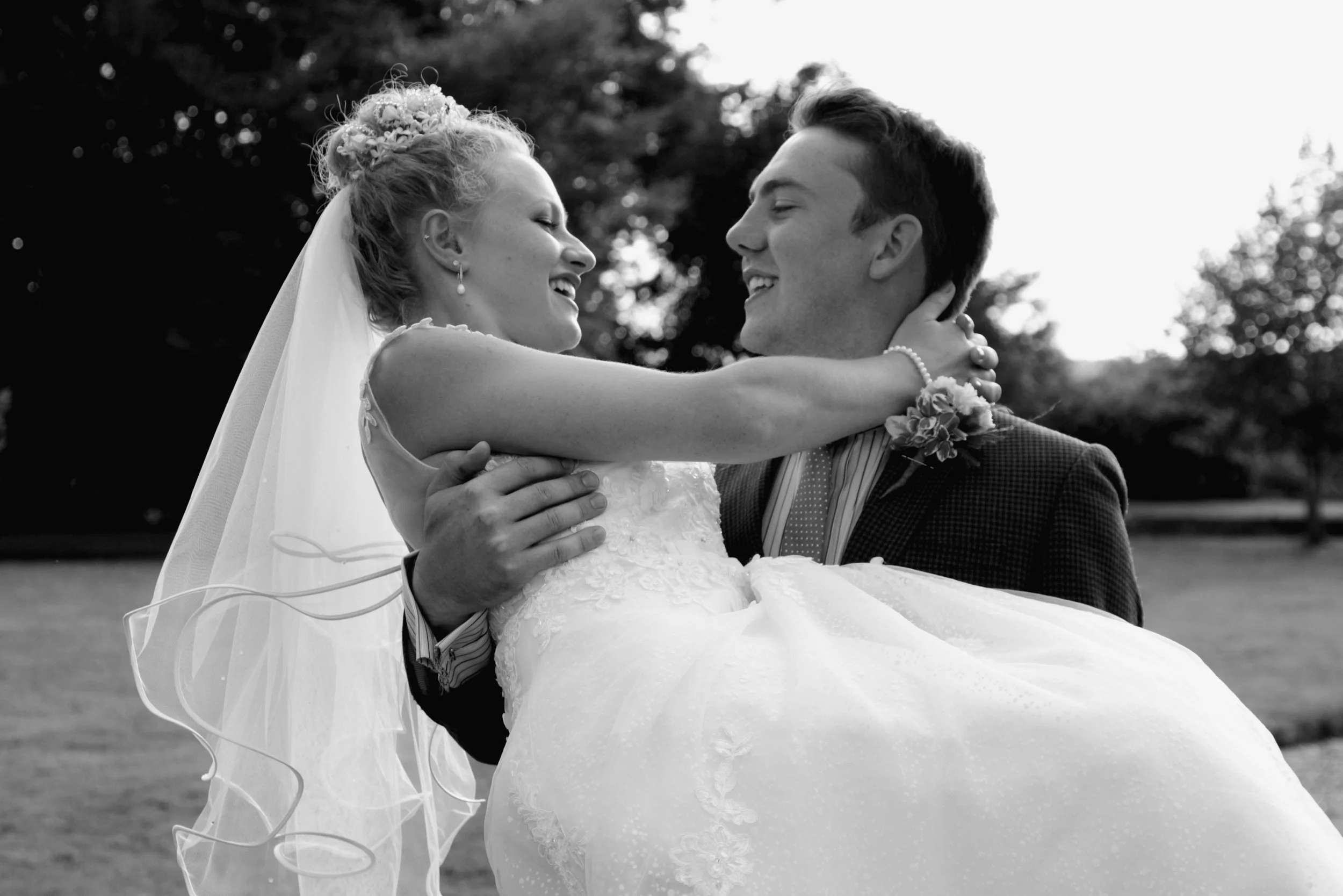 A bride and groom smiling and embracing outdoors, with trees in the background. The groom is carrying the bride, who is wearing a wedding dress and veil, with her arms around his neck.