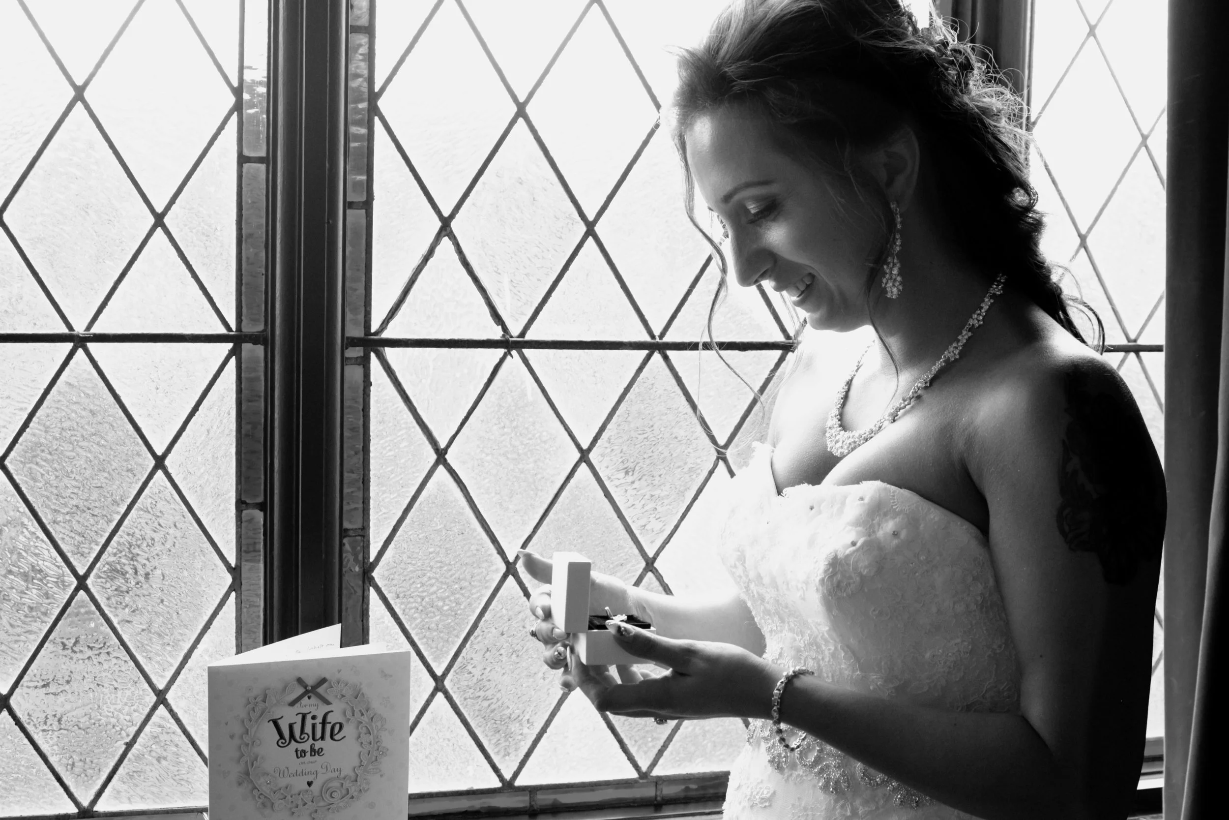 A bride in a strapless wedding gown standing near a window with diamond-shaped patterns, looking at a gift with a smile.