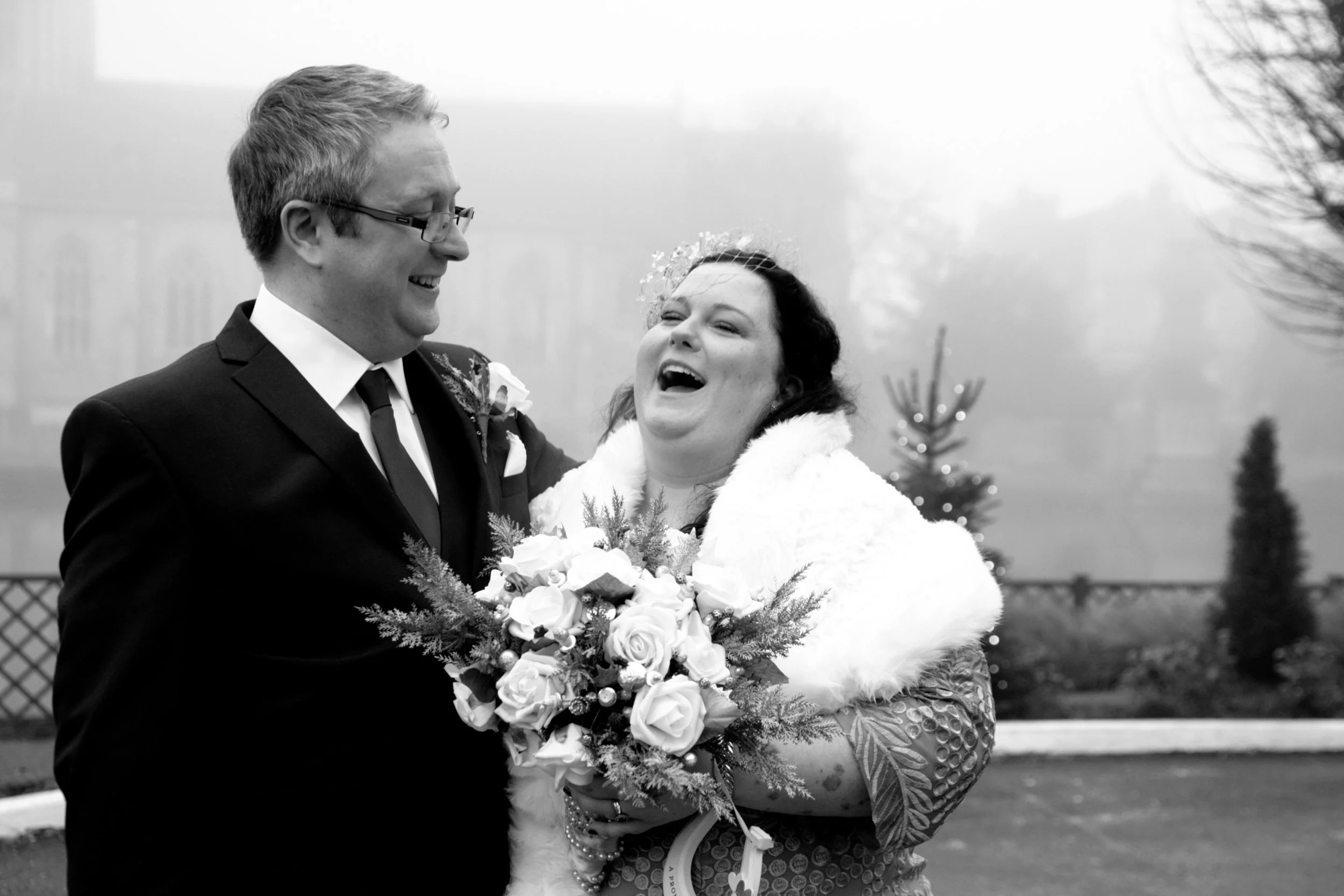 A groom in glasses and a suit laughs with a bride in a dress and a fur stole, holding a bouquet of flowers, on a foggy outdoor day.