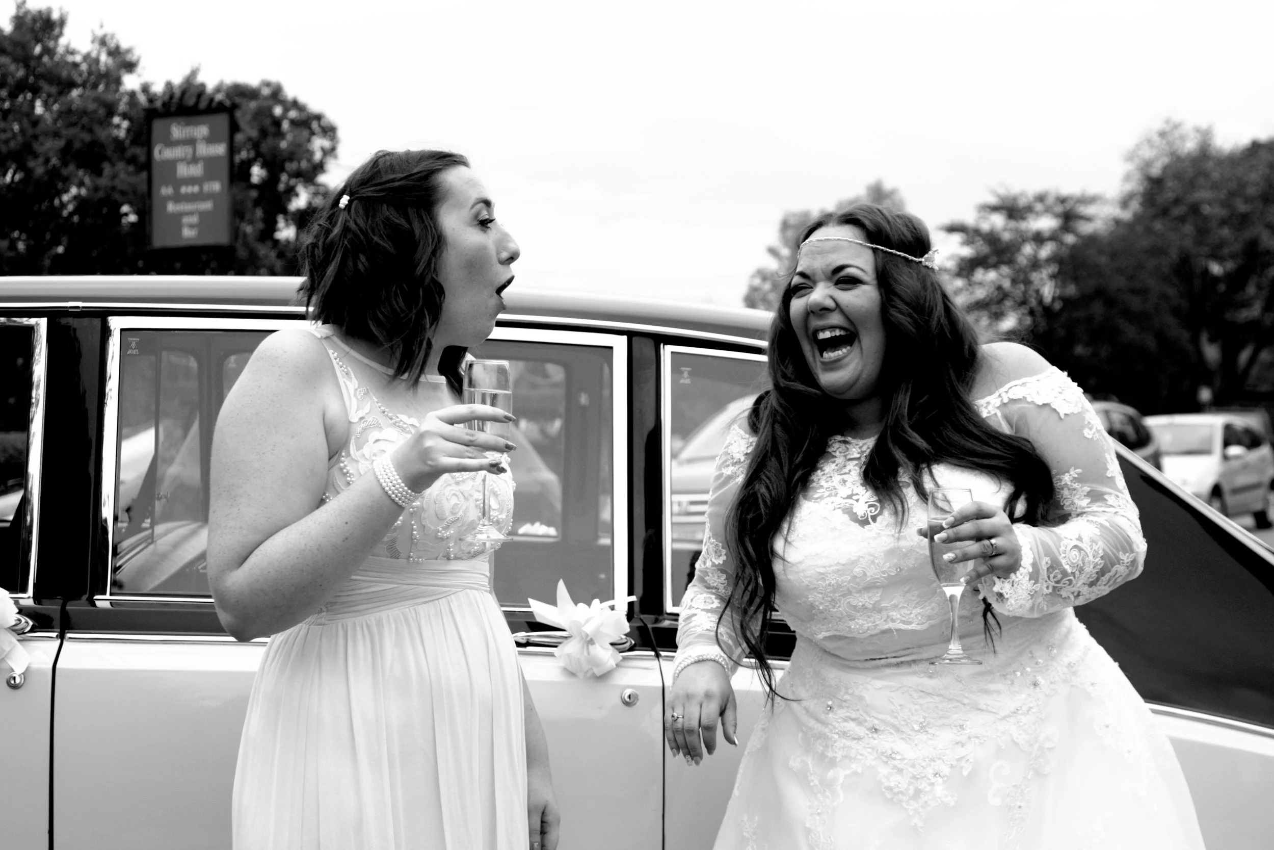 Two women in wedding dresses laughing and talking to each other while holding glasses of champagne,  standing in front of a vintage car.