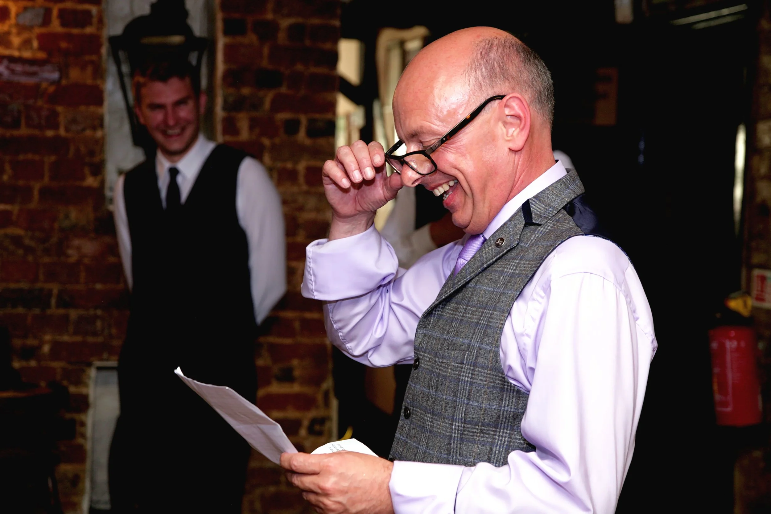 A father of the bride with glasses in a gray vest and pink shirt smiling while giving a speech, with his son-in-law in the background smiling, against a brick wall in a warmly lit room.