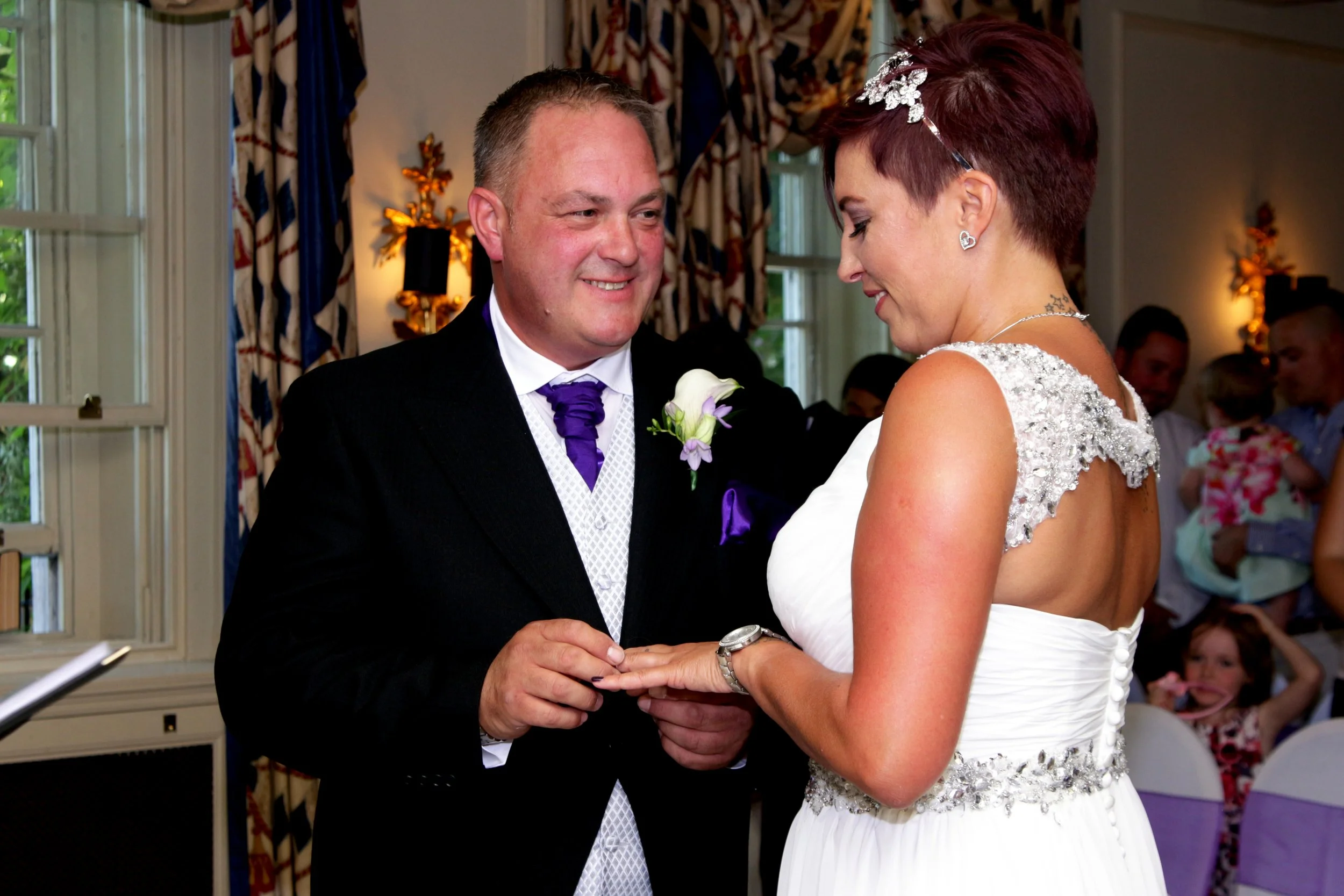 A bride and groom exchange wedding rings during their wedding ceremony indoors, with guests in the background.