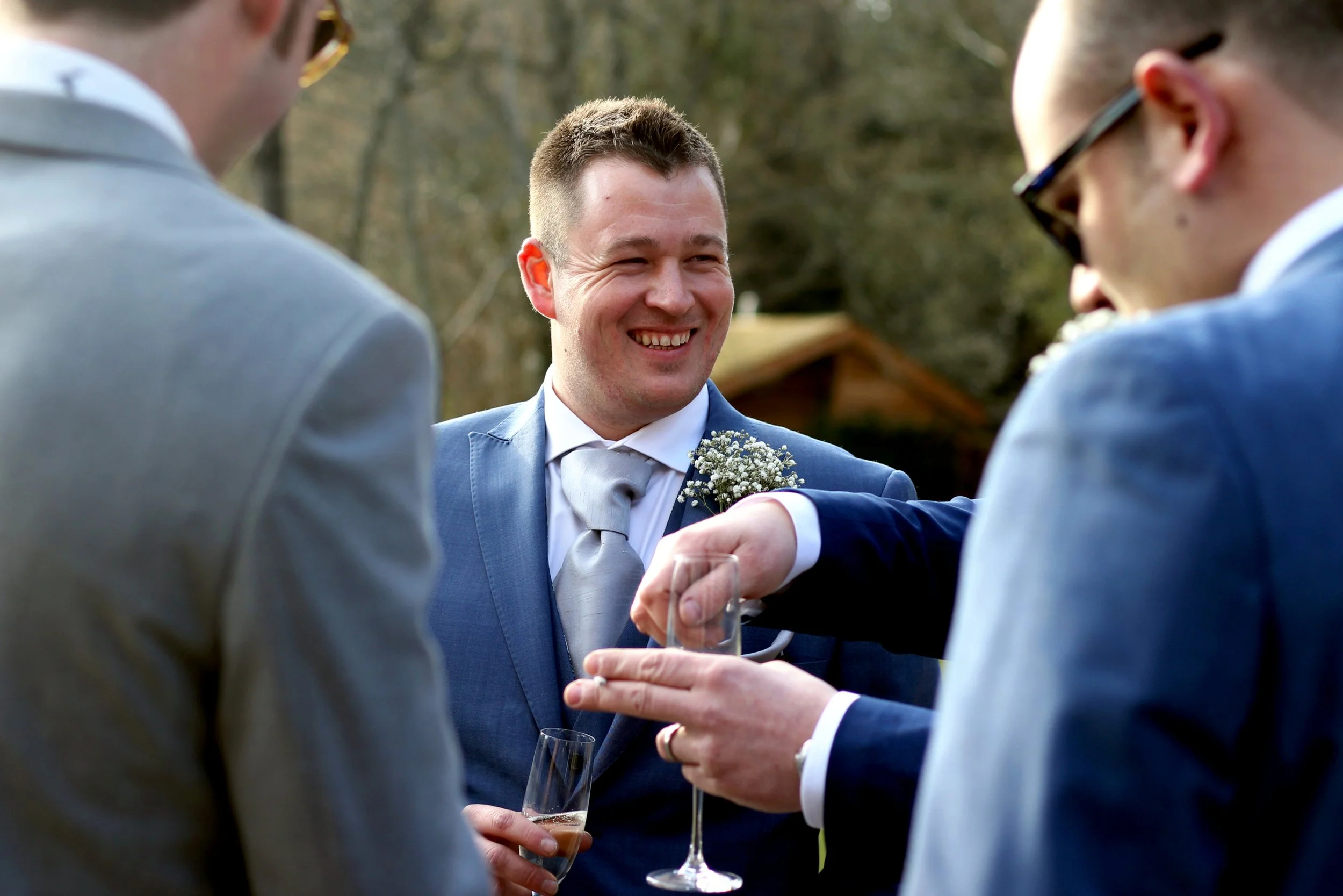 A groom in a blue suit and gray tie smiling and talking with three groomsmen at an outdoor wedding reception, holding glasses of champagne.