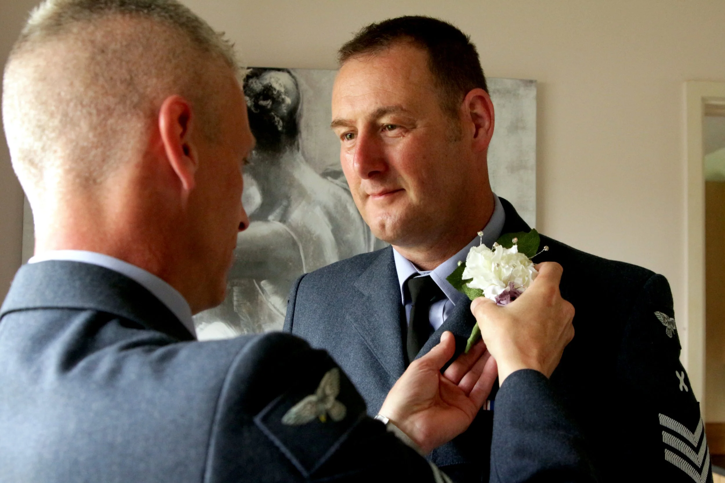 Two men in military uniforms, with one assisting the other by pinning on a white boutonniere, indoors.
