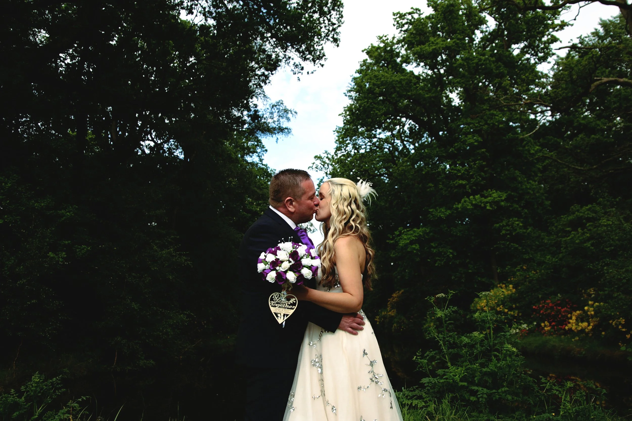 A newlywed couple sharing a kiss outdoors surrounded by trees, with the bride holding a bouquet of purple and white flowers.