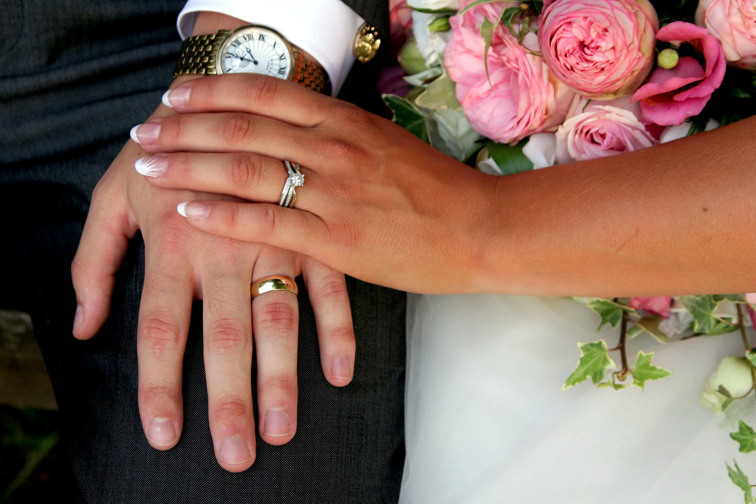 Close-up of a newly married couple's hands showing wedding bands and engagement ring, with a bouquet of pink roses and green leaves nearby.