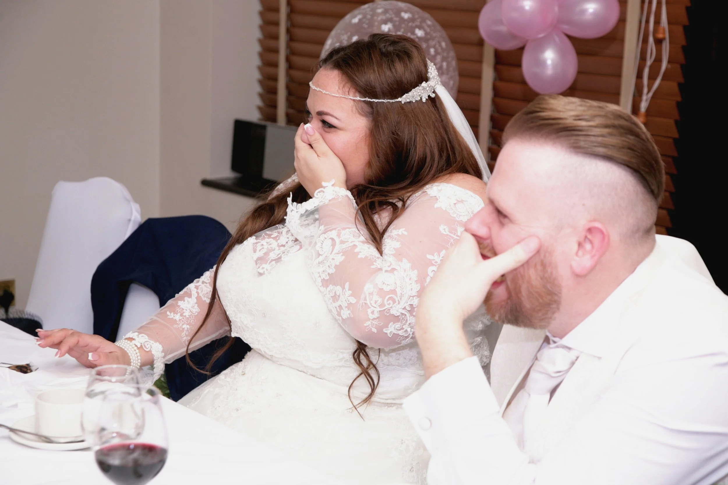 A bride and groom at their wedding reception, sharing a humorous moment, both laughing and covering their faces