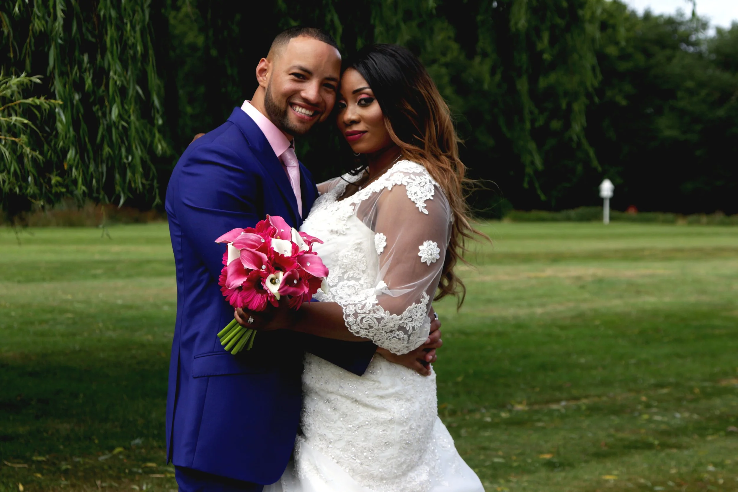 A newlywed couple embracing outdoors on a green lawn, with the groom wearing a blue suit and pink shirt, the bride wearing a white lace wedding gown with sheer sleeves and holding a bouquet of pink and white flowers.