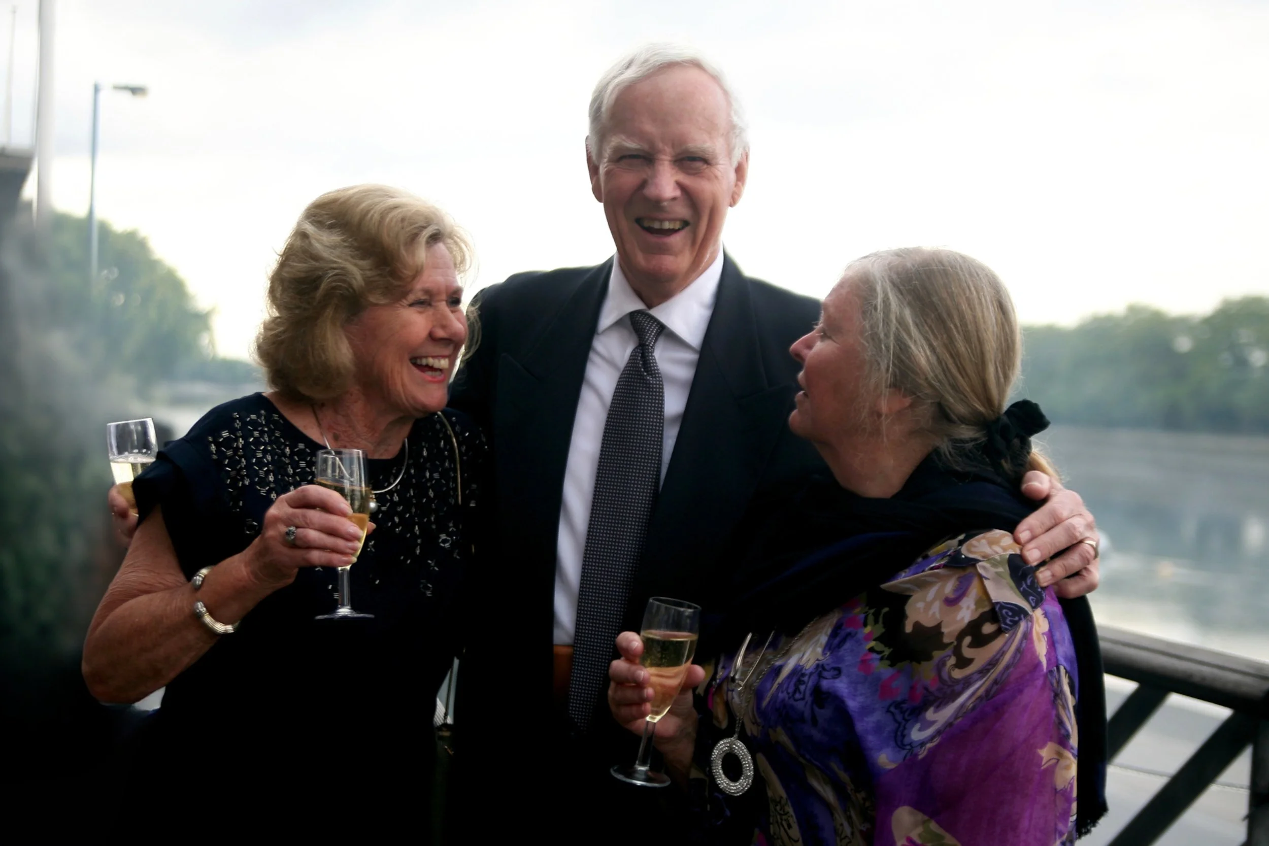 Three older adults celebrating a wedding with glasses of champagne outdoors, smiling and embracing.