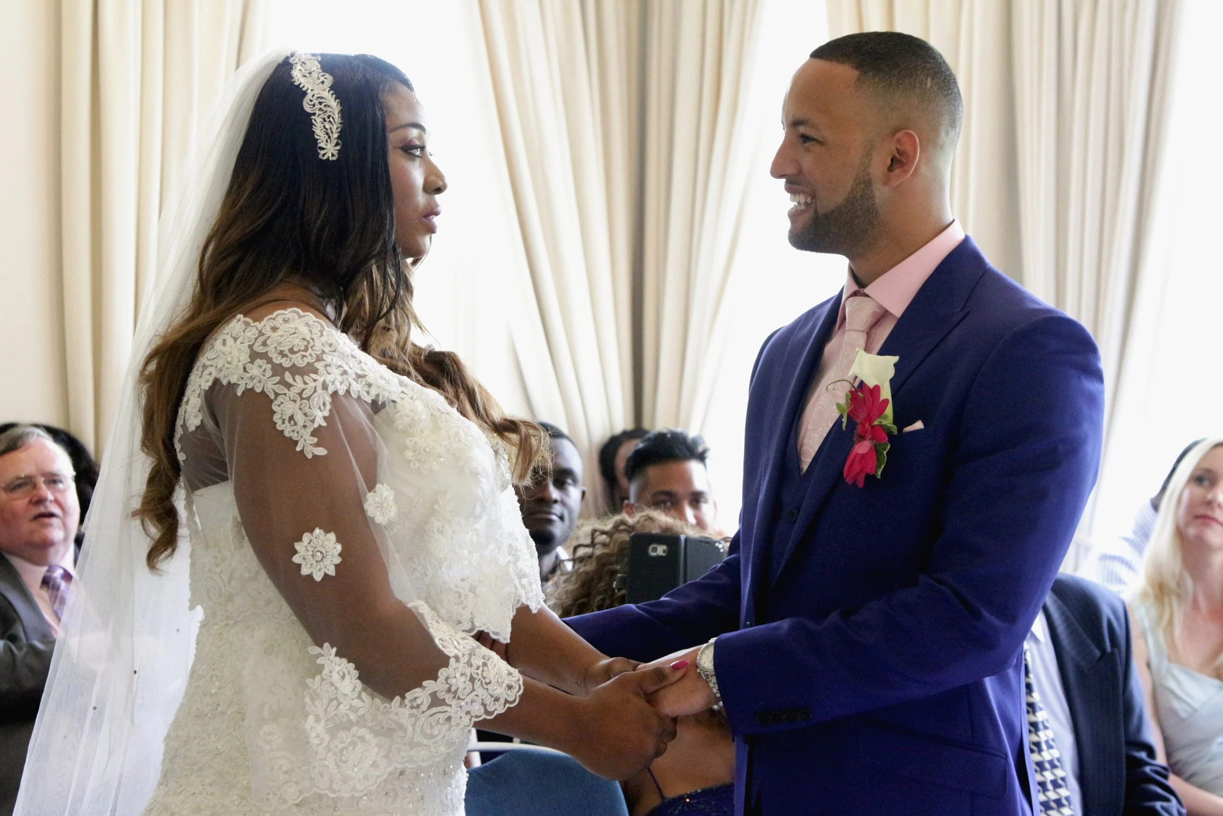 A bride and groom holding hands during wedding ceremony, standing indoors with seated guests in the background.