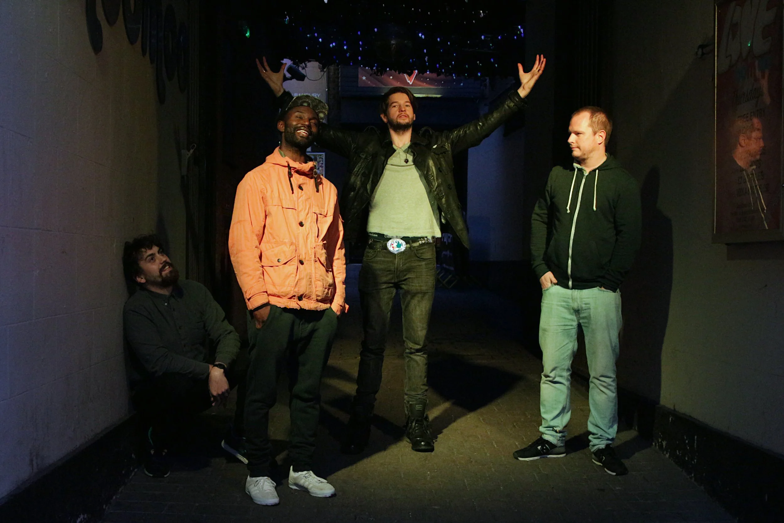 Five men in a dimly lit hallway, with one standing in the center with arms raised and four others near him, some standing and one squatting, engaging in casual poses.
