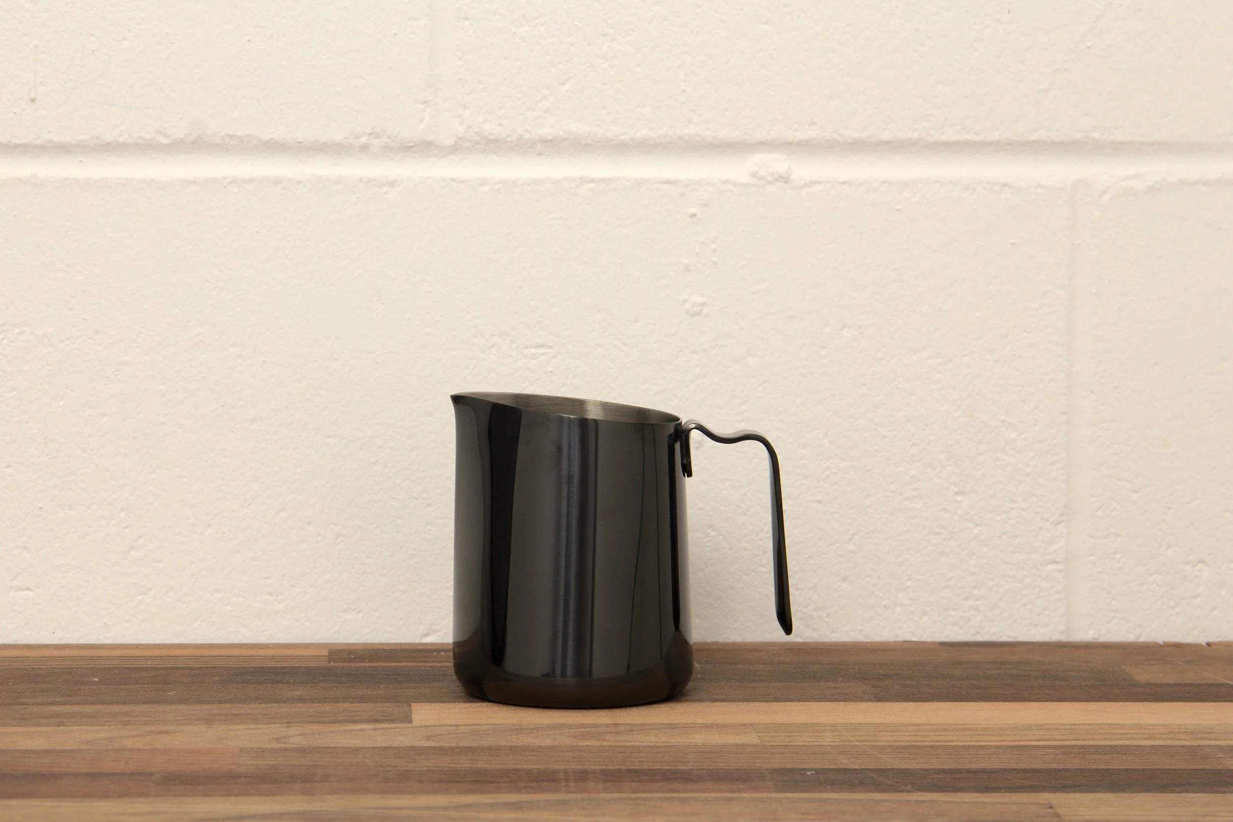 A black milk pitcher with a handle on a wooden surface against a white brick wall background.