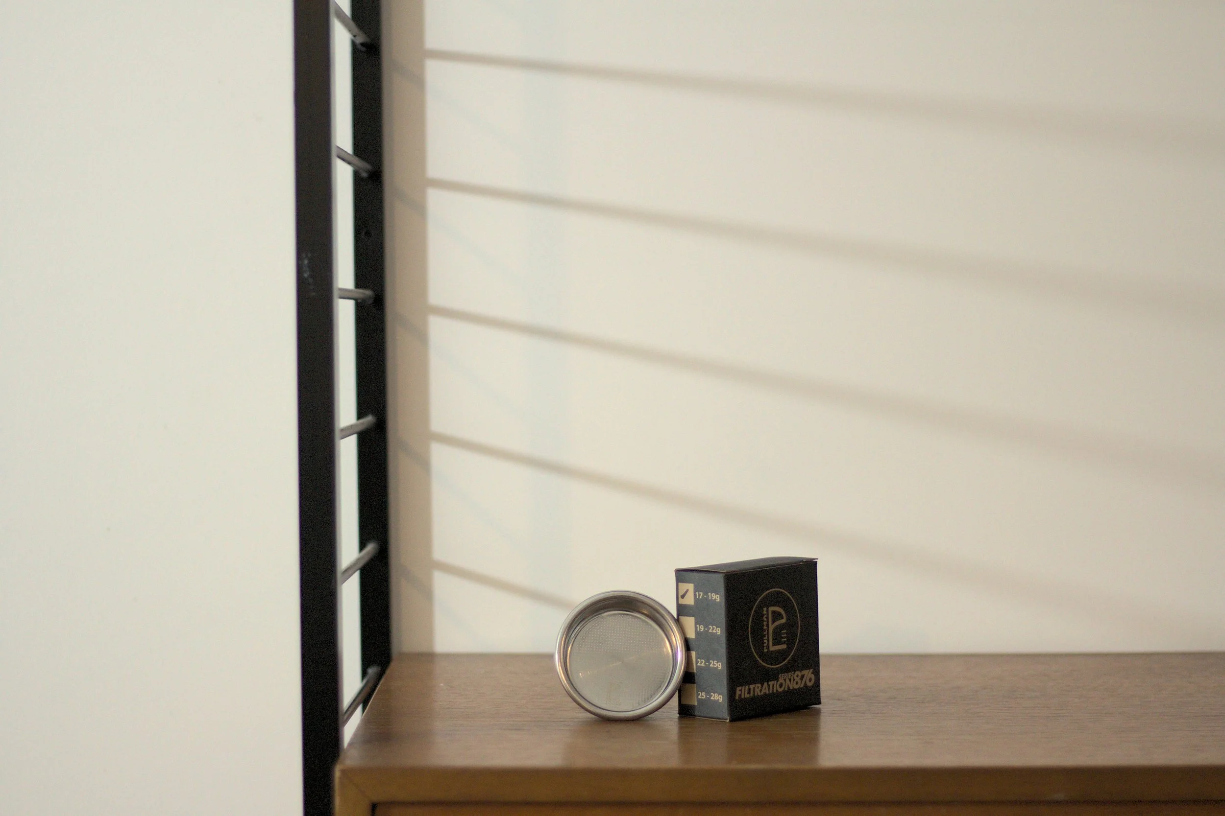 A silver portafilter basket and black box on a wooden shelf with a white wall and shadows in the background.
