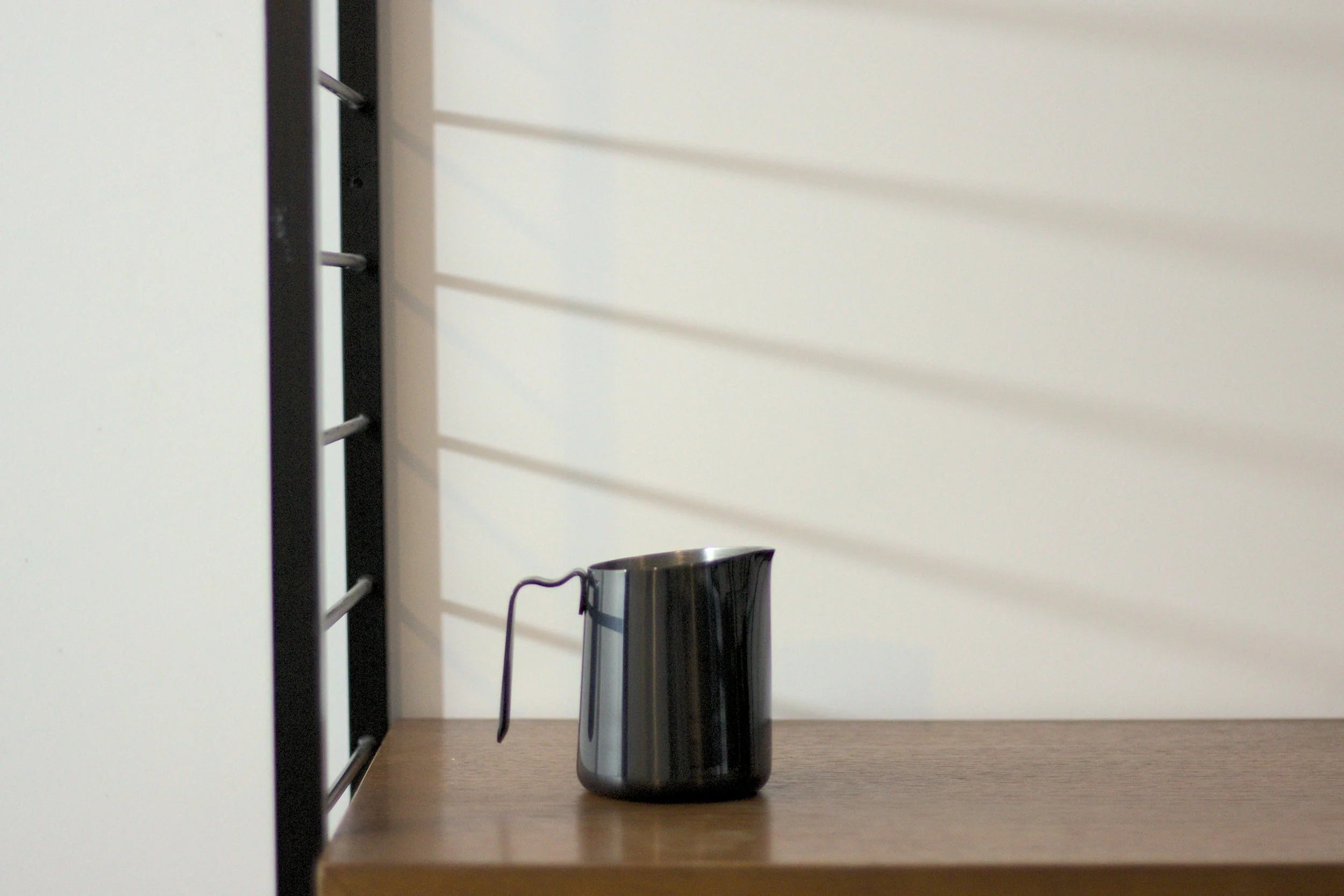 A black milk pitcher with a handle sitting on a wooden shelf against a white wall with shadows