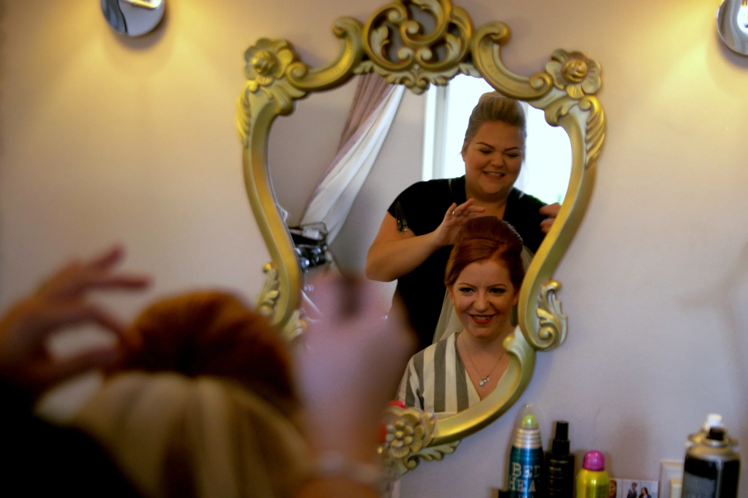A woman with red hair getting ready, looking into a decorative mirror as a hairstylist styles her hair. The mirror reflects her smiling face, and salon products are on the counter below.