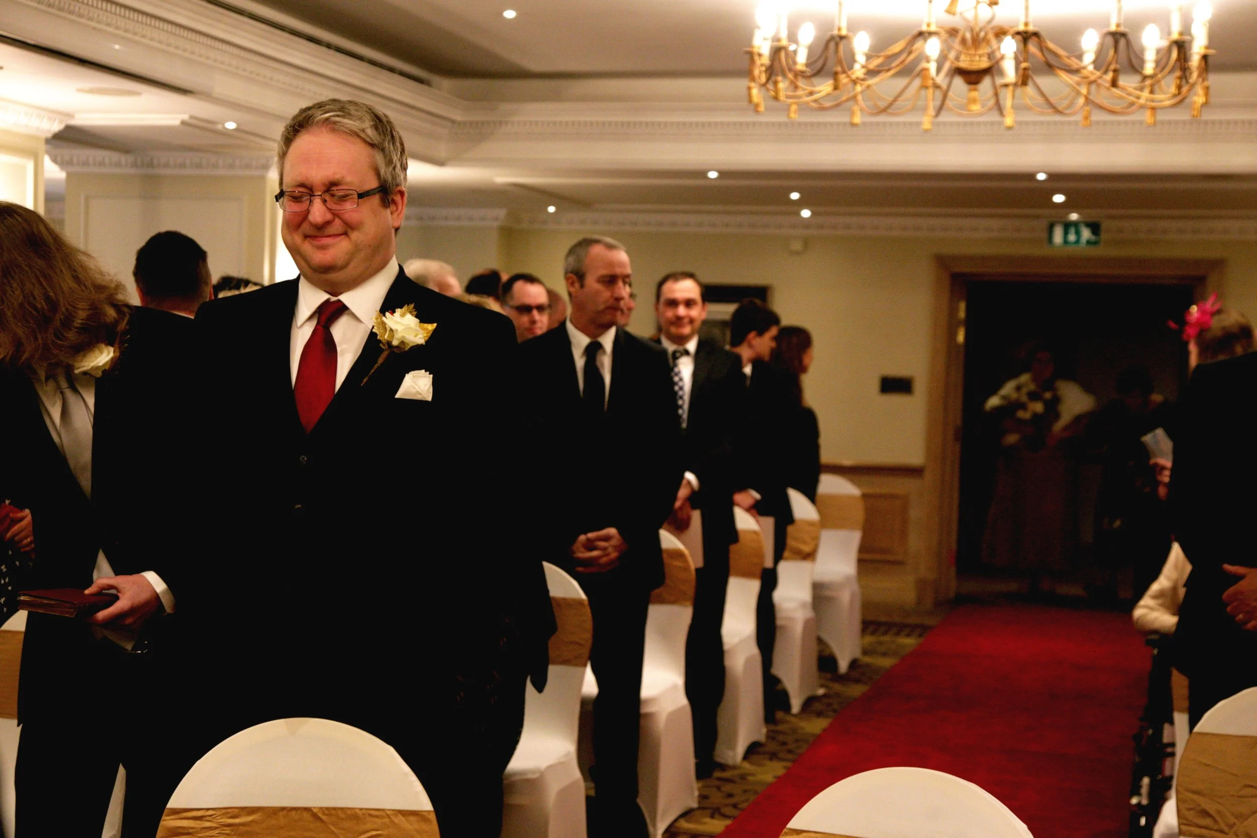 A groom in glasses, a suit, and a red tie, waiting for his bride to walk down the aisle in a decorated indoor venue featuring ornate chandeliers and white chairs with gold sashes.