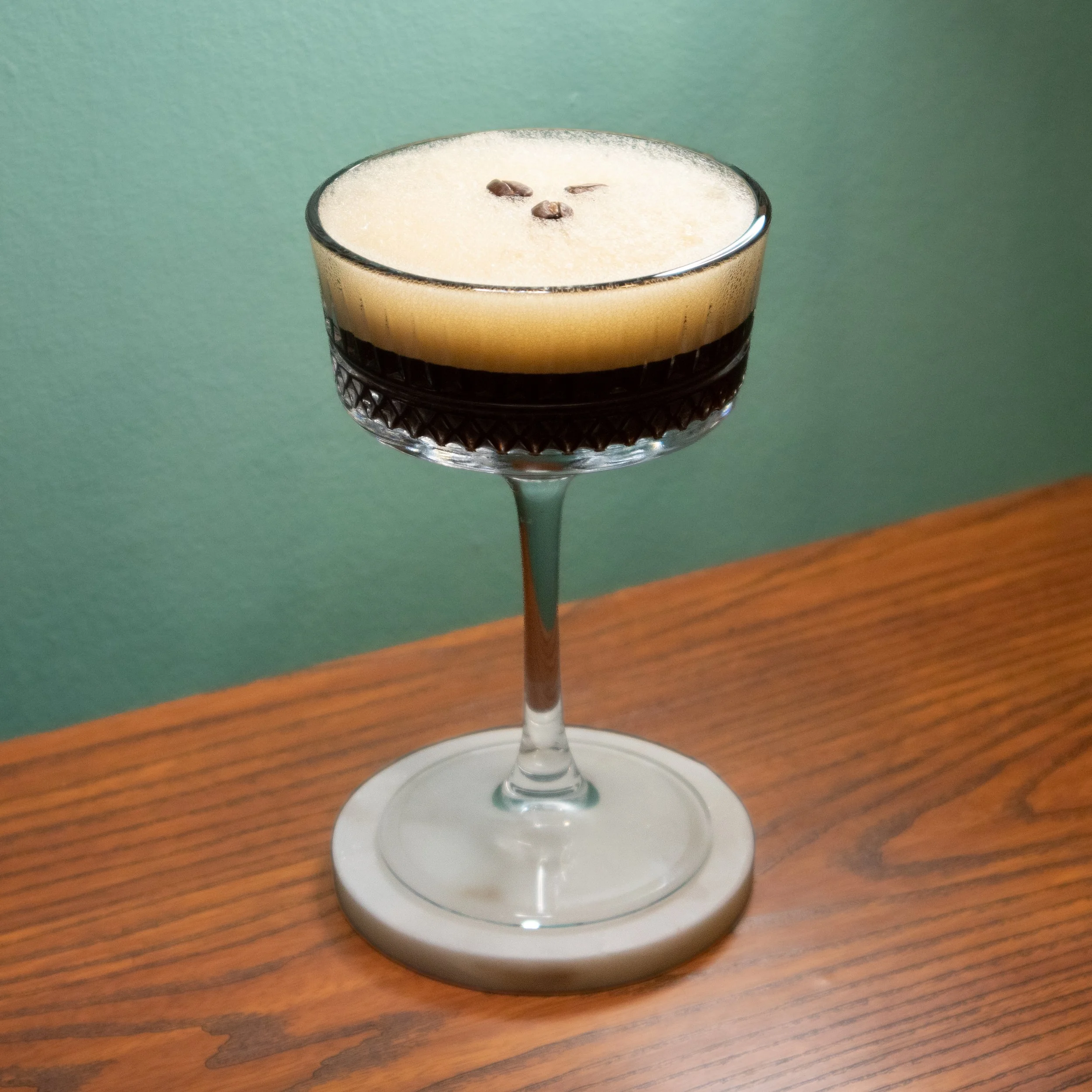 A cocktail glass with a dark beverage topped with frothy foam and three small coffee beans, placed on a white decorative coaster on a wooden surface with a green wall in the background.