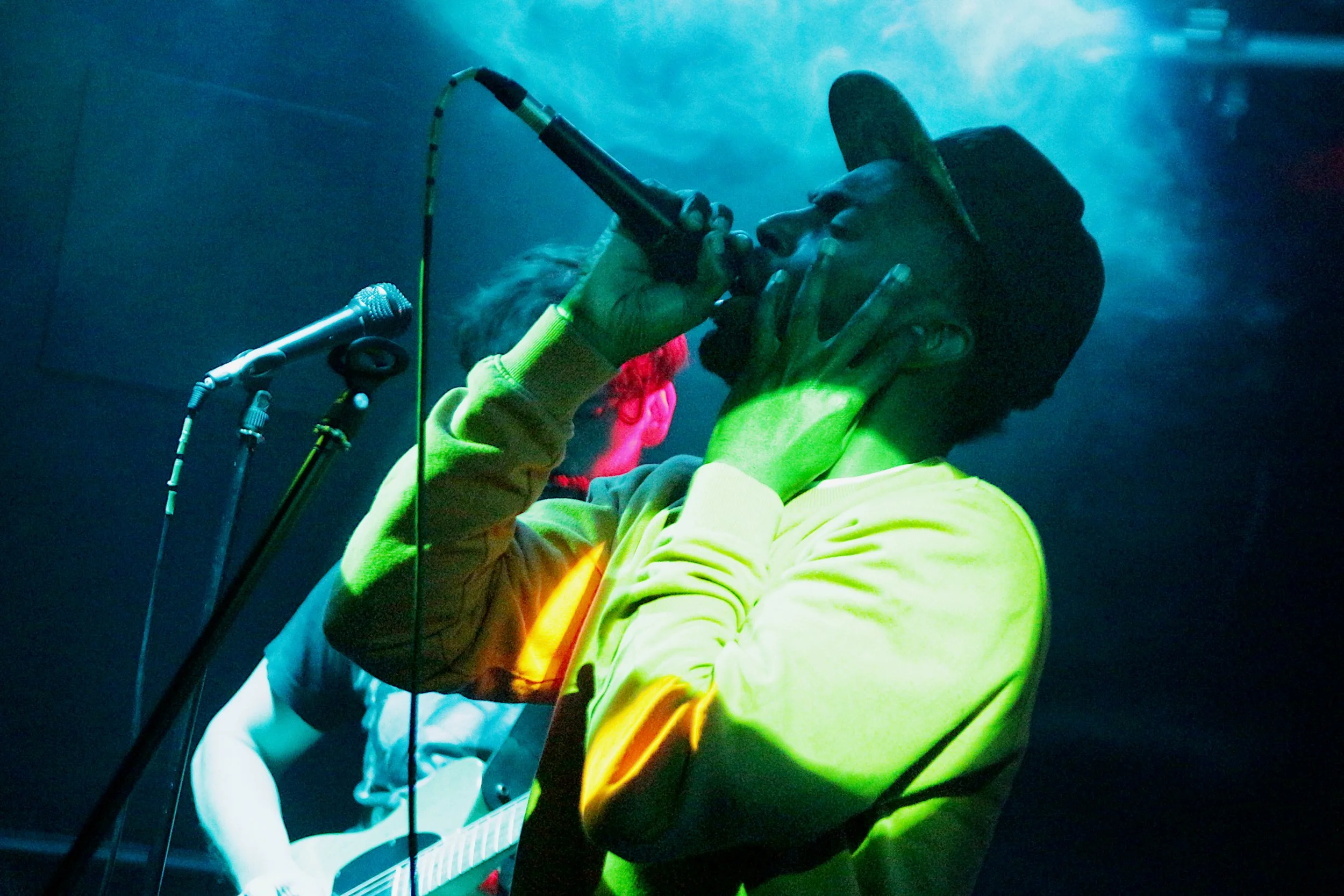 Two musicians perform on stage, one singing into a microphone and the other playing the guitar, with blue and green lighting creating an energetic atmosphere.
