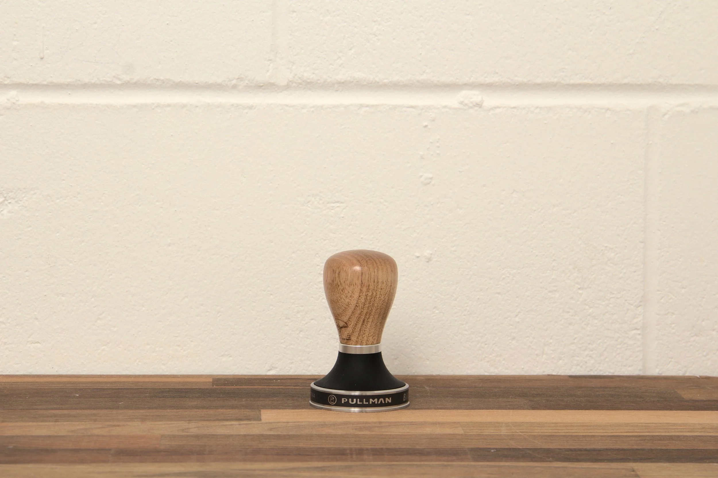 A wooden-handled coffee tamper on a wooden surface against a white brick wall background. 
