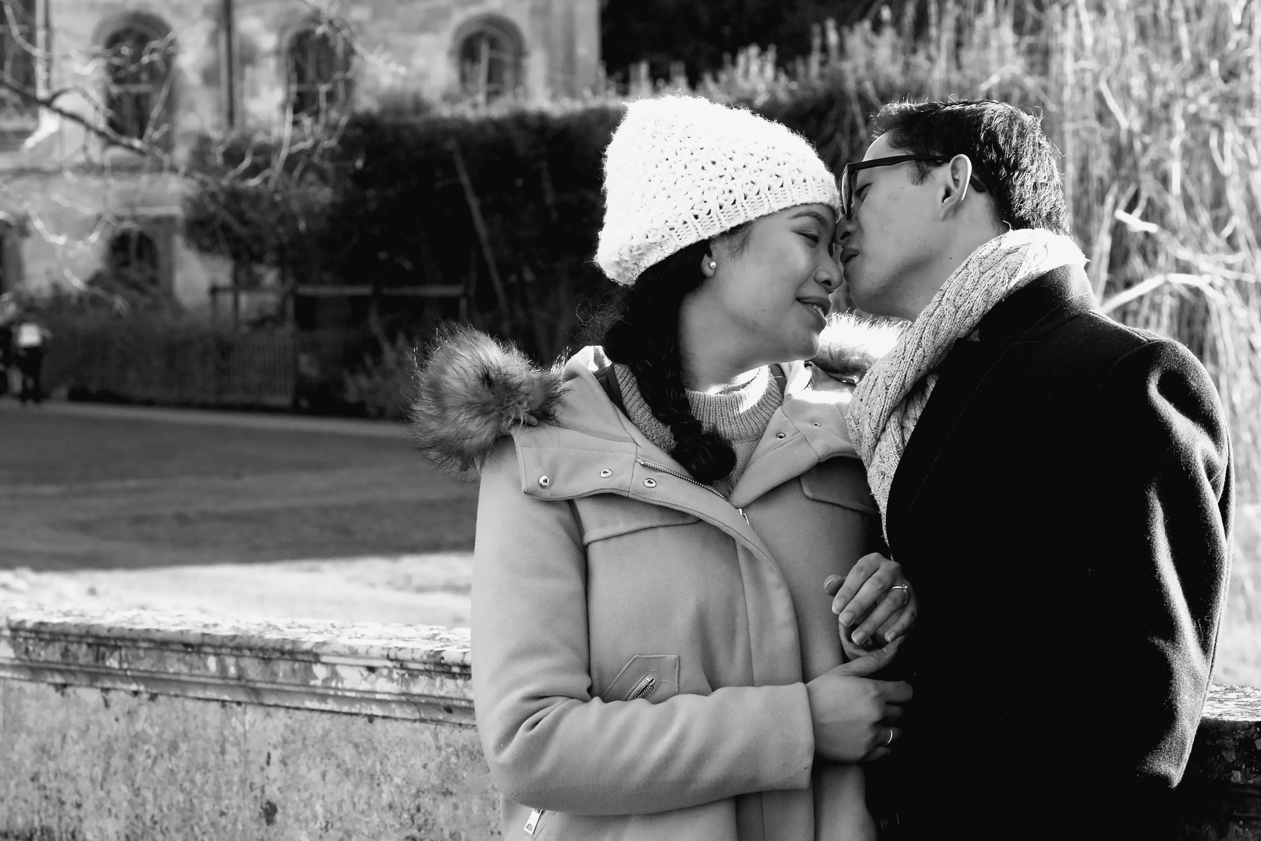 A couple standing close together outdoors, with their foreheads touching, smiling and sharing a tender moment. The woman wears a knit beanie and winter coat with a fur-lined hood, while the man wears glasses, a scarf, and a coat.