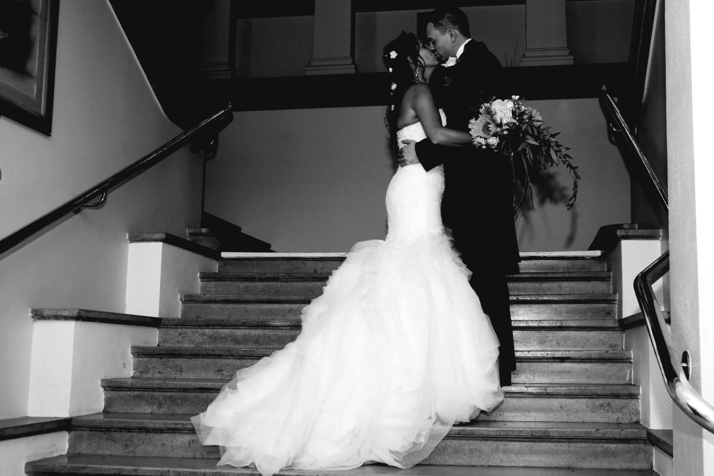 A newlywed couple sharing a kiss on a staircase, the bride in a white wedding gown with a long train and the groom in a black tuxedo, the bride holding a bouquet of flowers.
