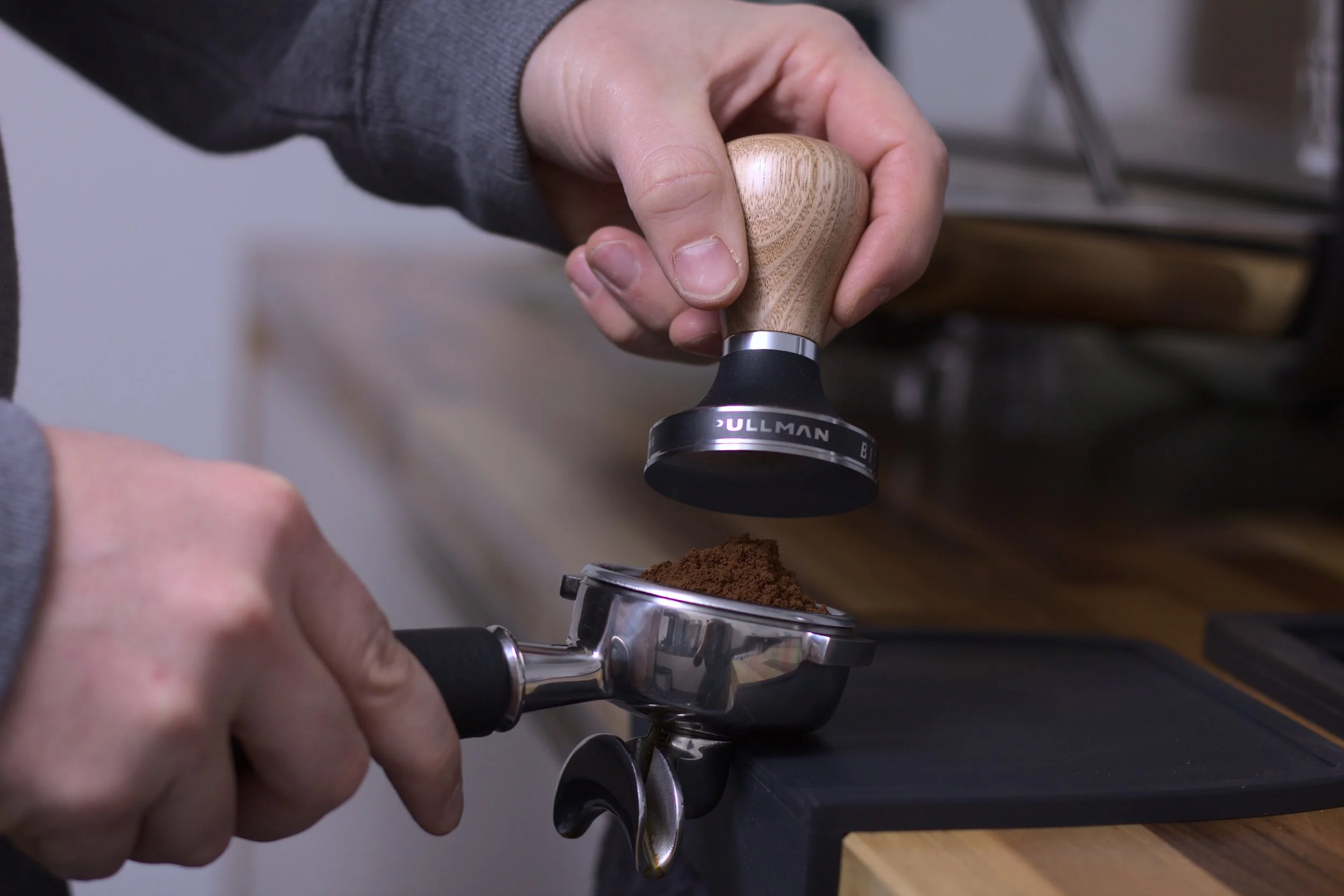 A person using a wood-handled tamper to press ground coffee into an espresso portafilter.