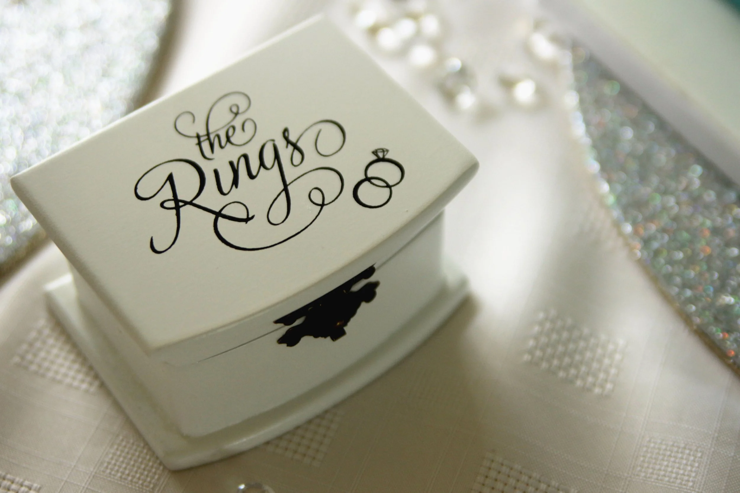 A white jewelry box with black cursive lettering that reads 'the Rungs' on the lid, placed on a white textured surface with glittery embellishments.