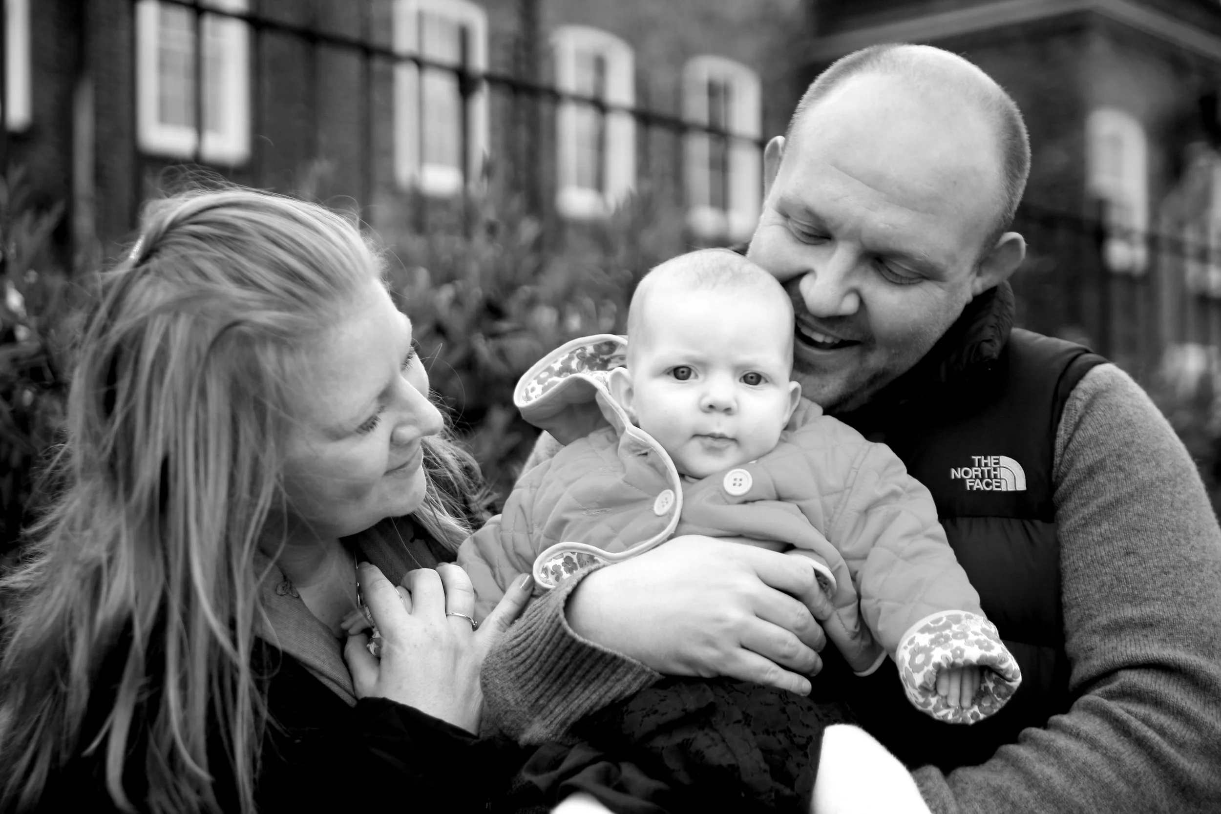A family of three, a woman, a man, and a baby, enjoying time outdoors. The man is holding the baby, who is wearing a jacket, while the woman looks at them smiling.