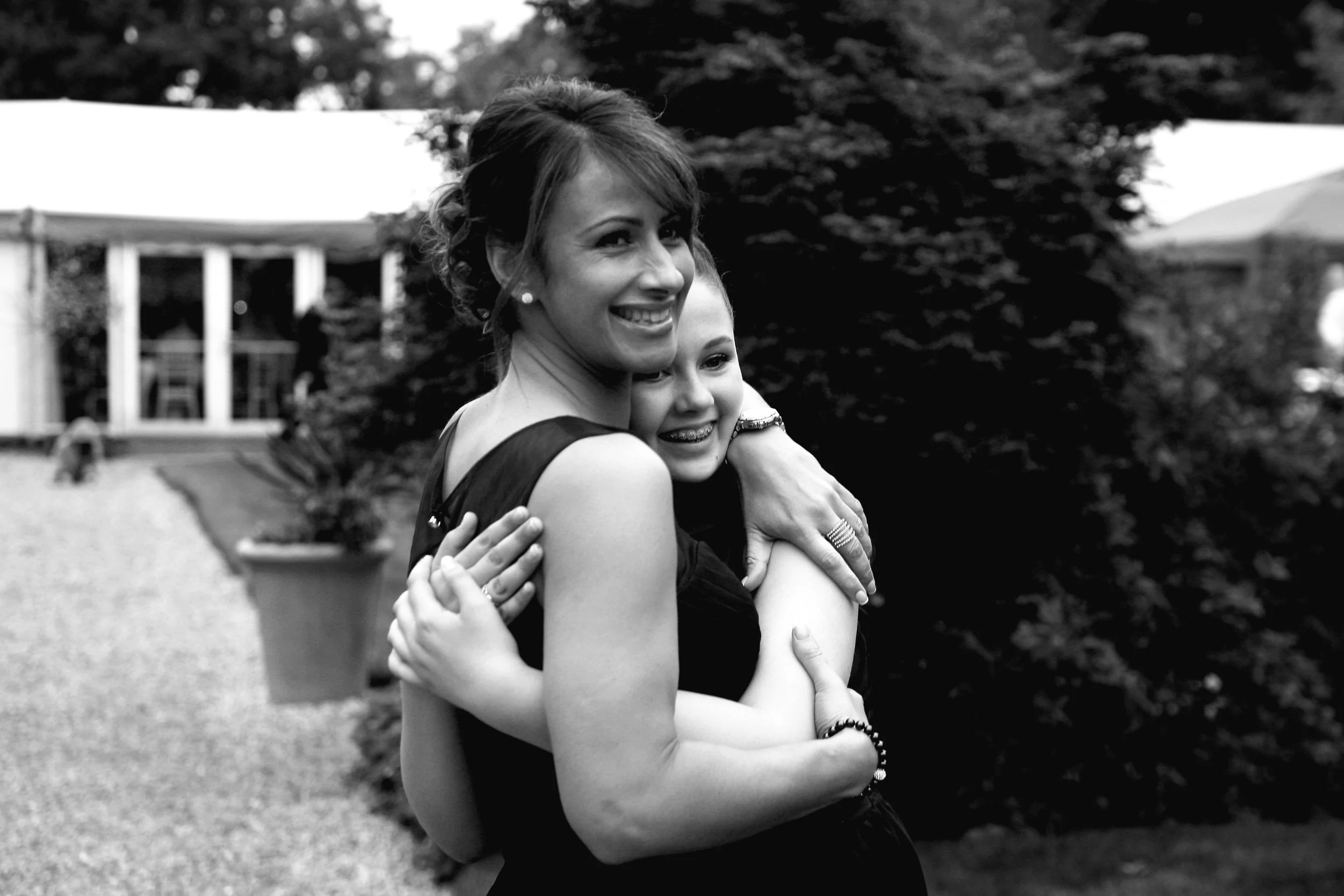 A pair of bridesmaids, a woman and a young girl, hugging outdoors, both smiling. There are trees and a building in the background.