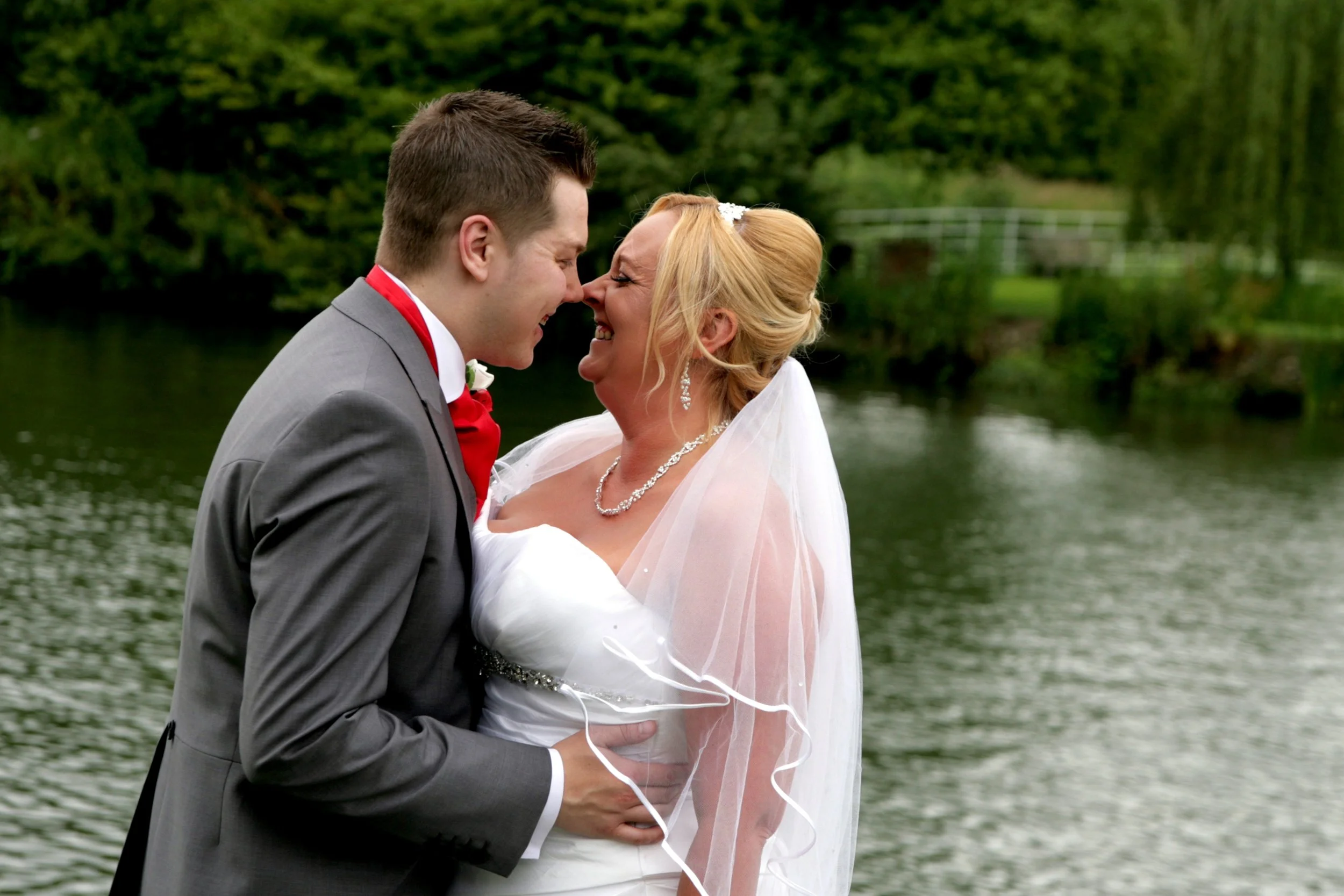 A bride and groom smiling and touching foreheads by a lake, with trees and a bridge in the background.