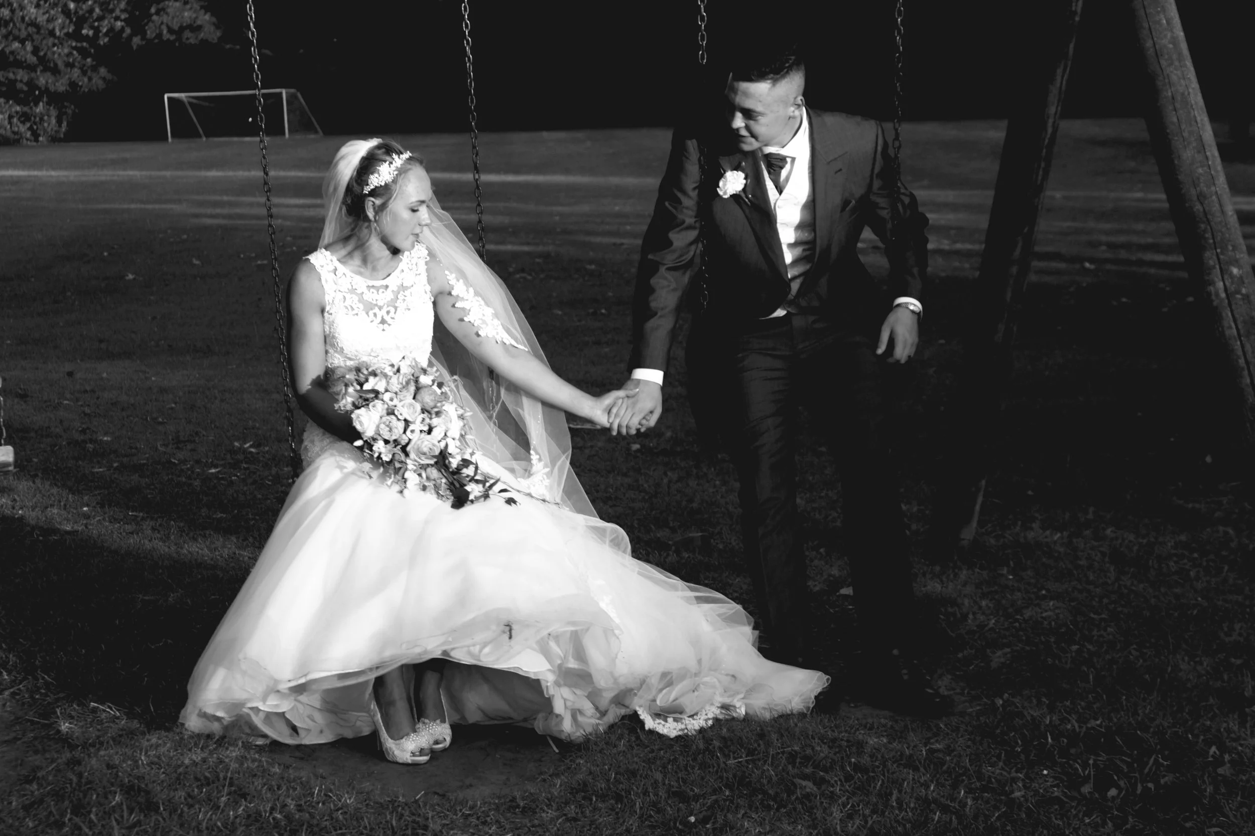 A bride and groom sitting on a swing in a park at night, holding hands, with the bride holding a bouquet of flowers.