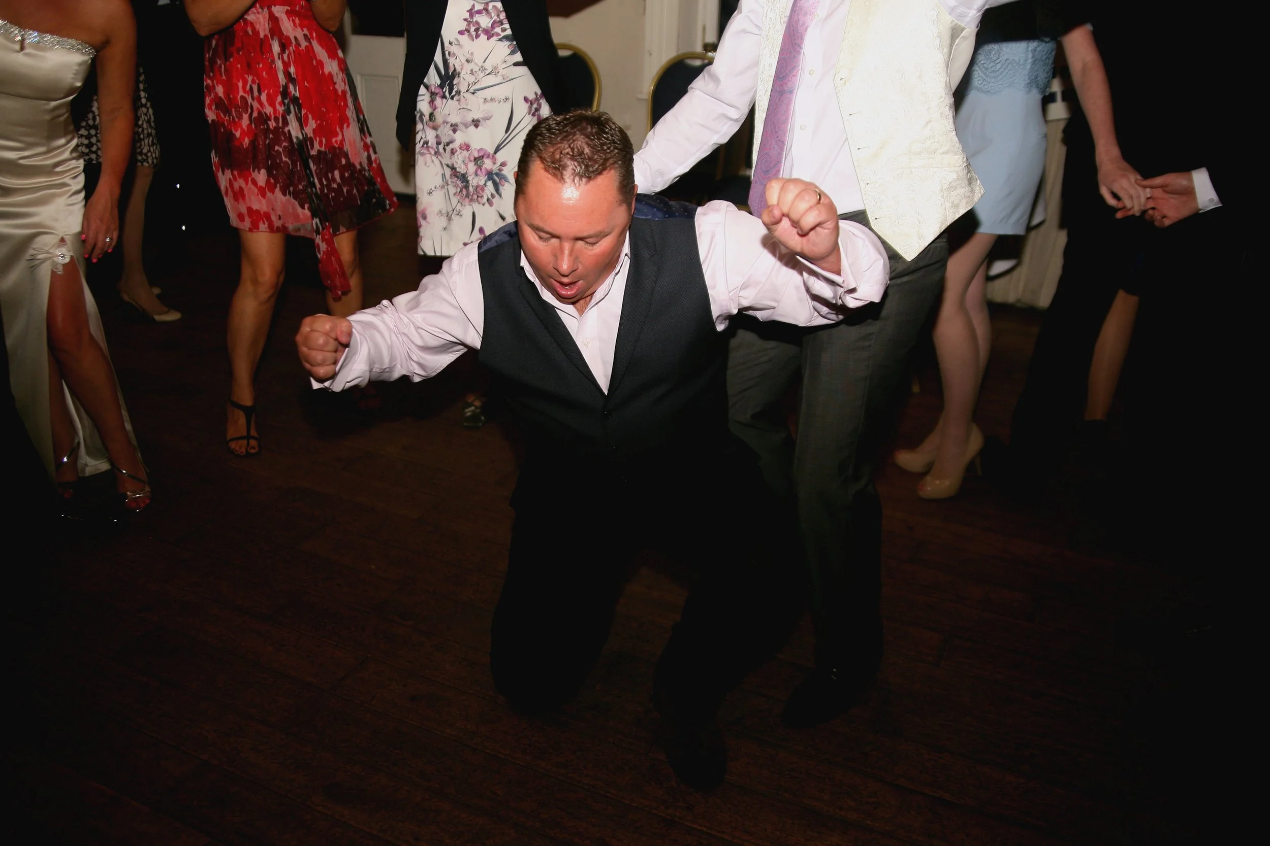 A man in a tuxedo kneeling on the dance floor with his arms raised, appearing to dance or celebrate at a social event surrounded by other people in formal attire.