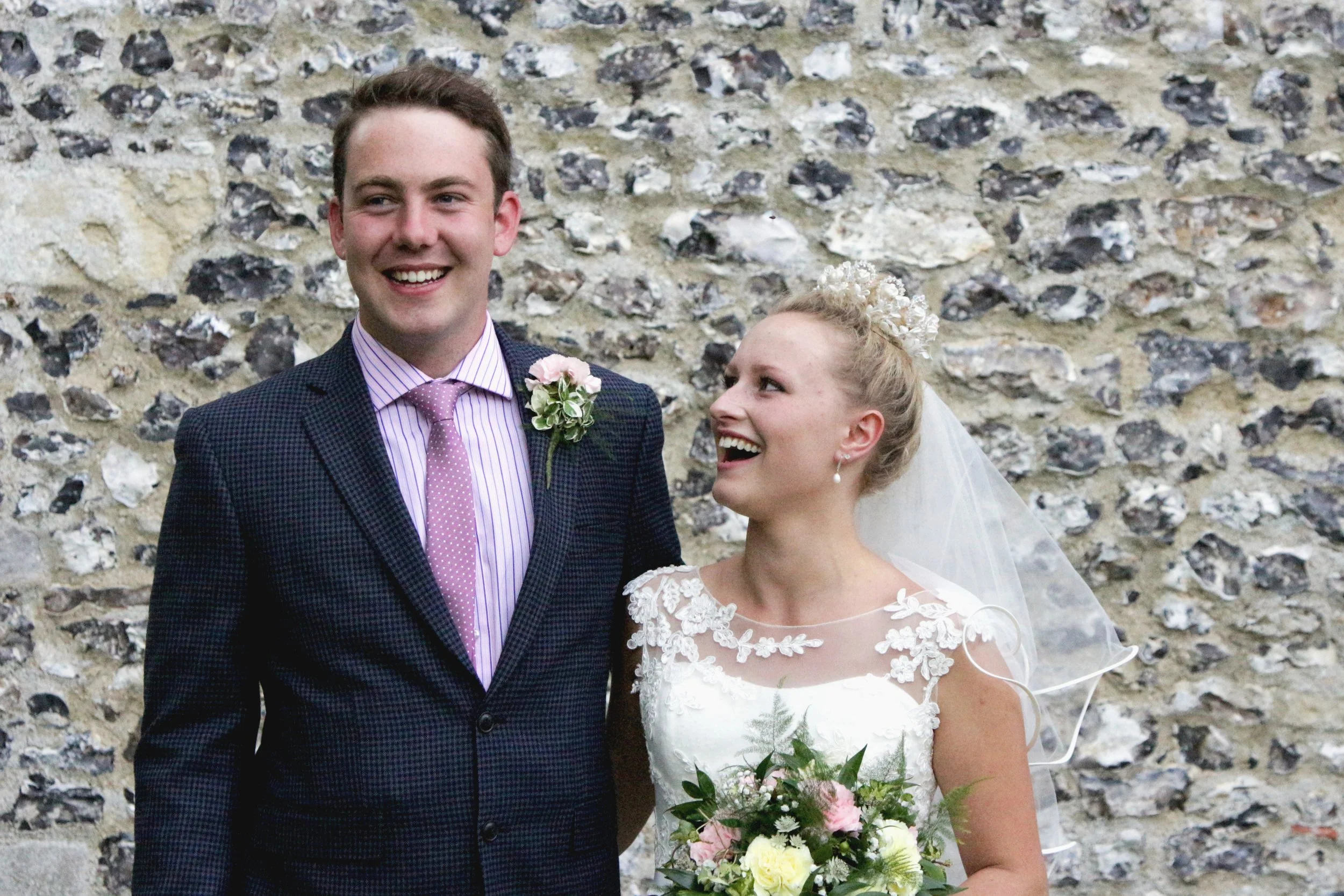 A smiling bride and groom stand together in front of a stone wall, with the bride holding a bouquet of pink and white flowers, and wearing a wedding dress with lace details and a veil, while the groom wears a patterned suit and a pink striped tie.
