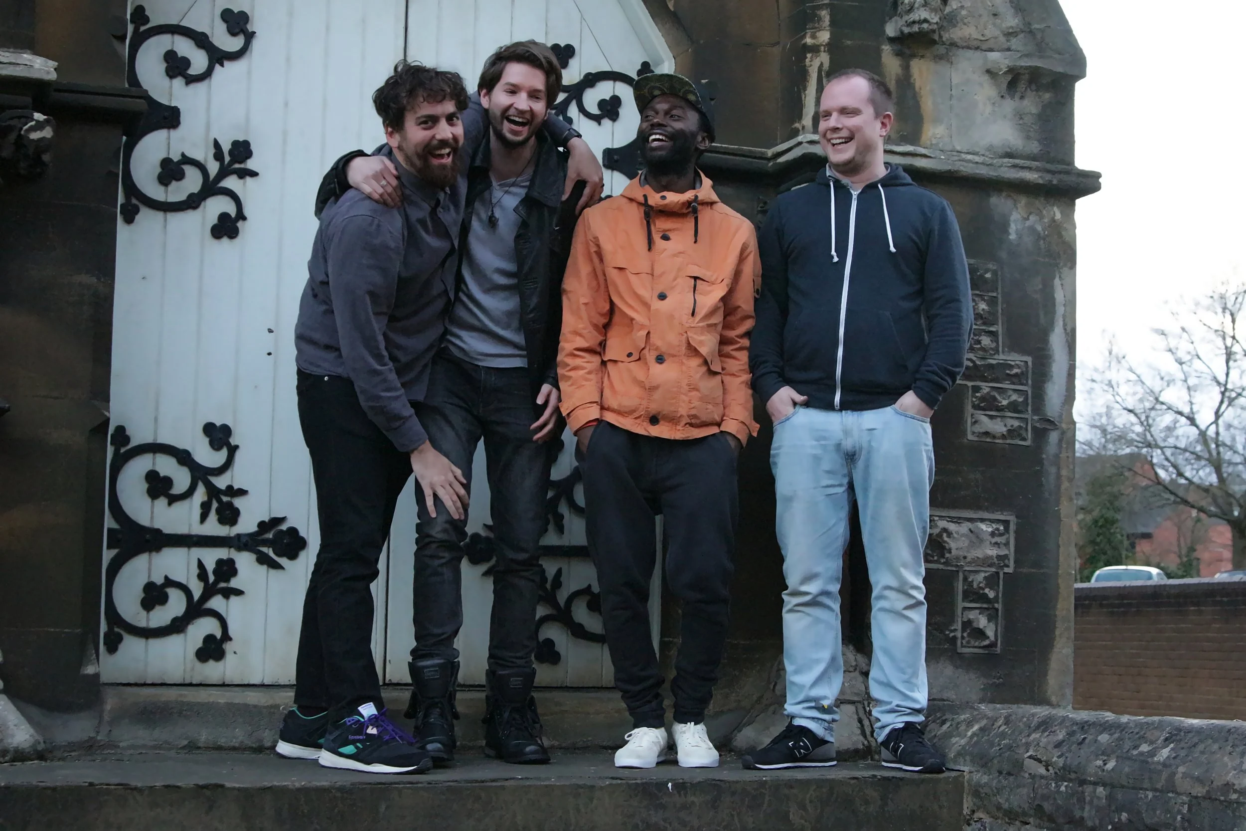 Four men standing outside near a stone wall and a white door with black decorative ironwork, smiling and enjoying each other's company.