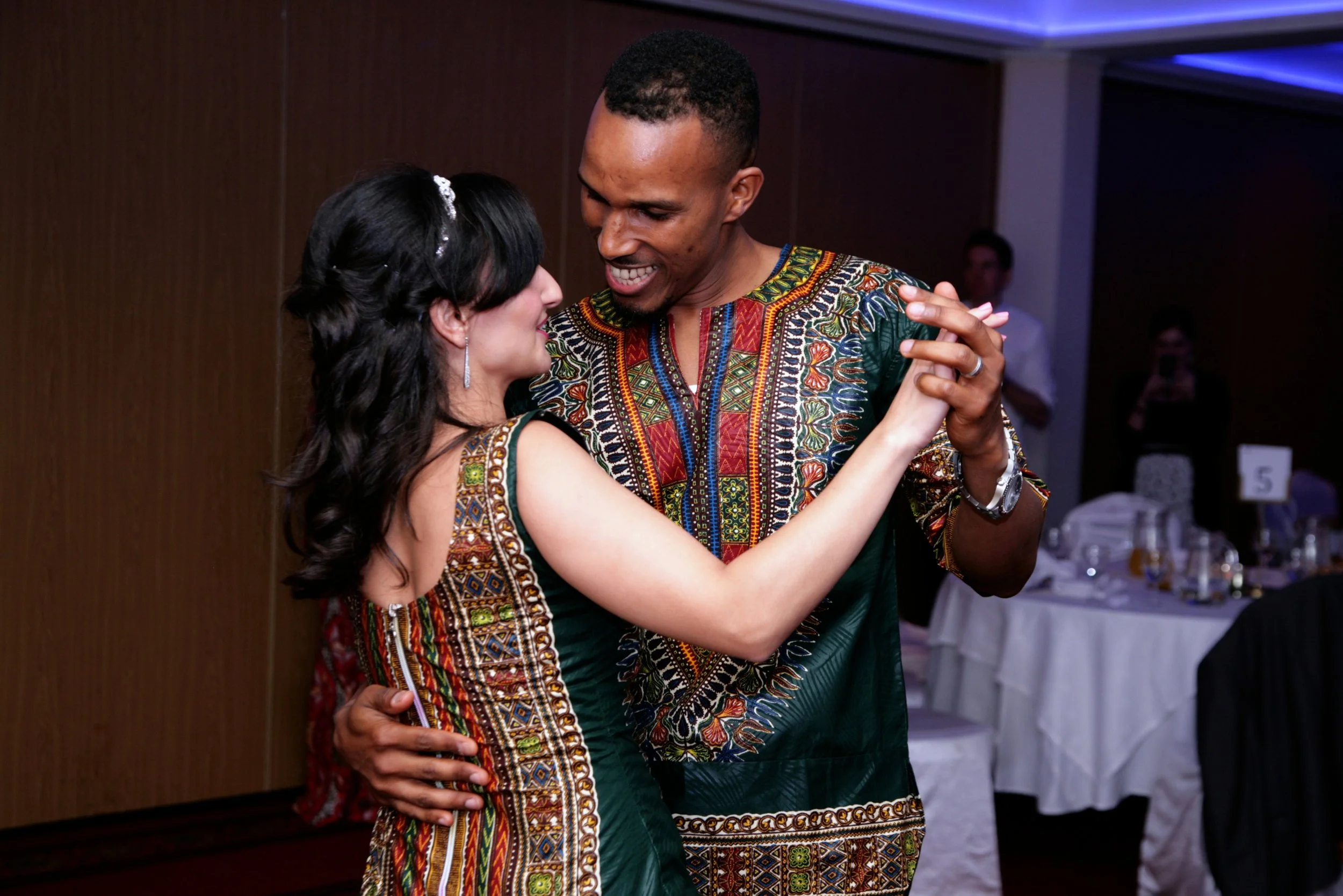 A bride and groom dancing together at an indoor event, dressed in colorful, patterned clothing, smiling and enjoying the moment.