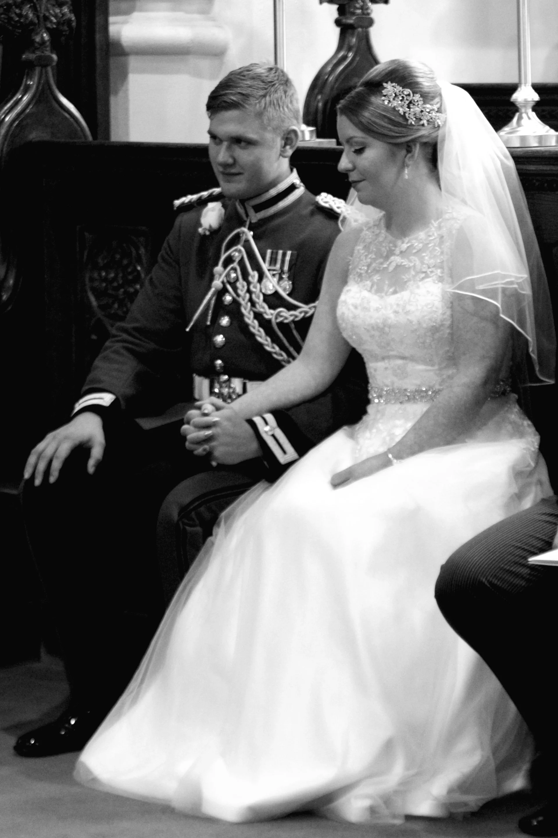A bride and groom sitting together at their wedding ceremony, holding hands. The groom is dressed in a military uniform, and the bride is wearing a wedding dress with a veil.