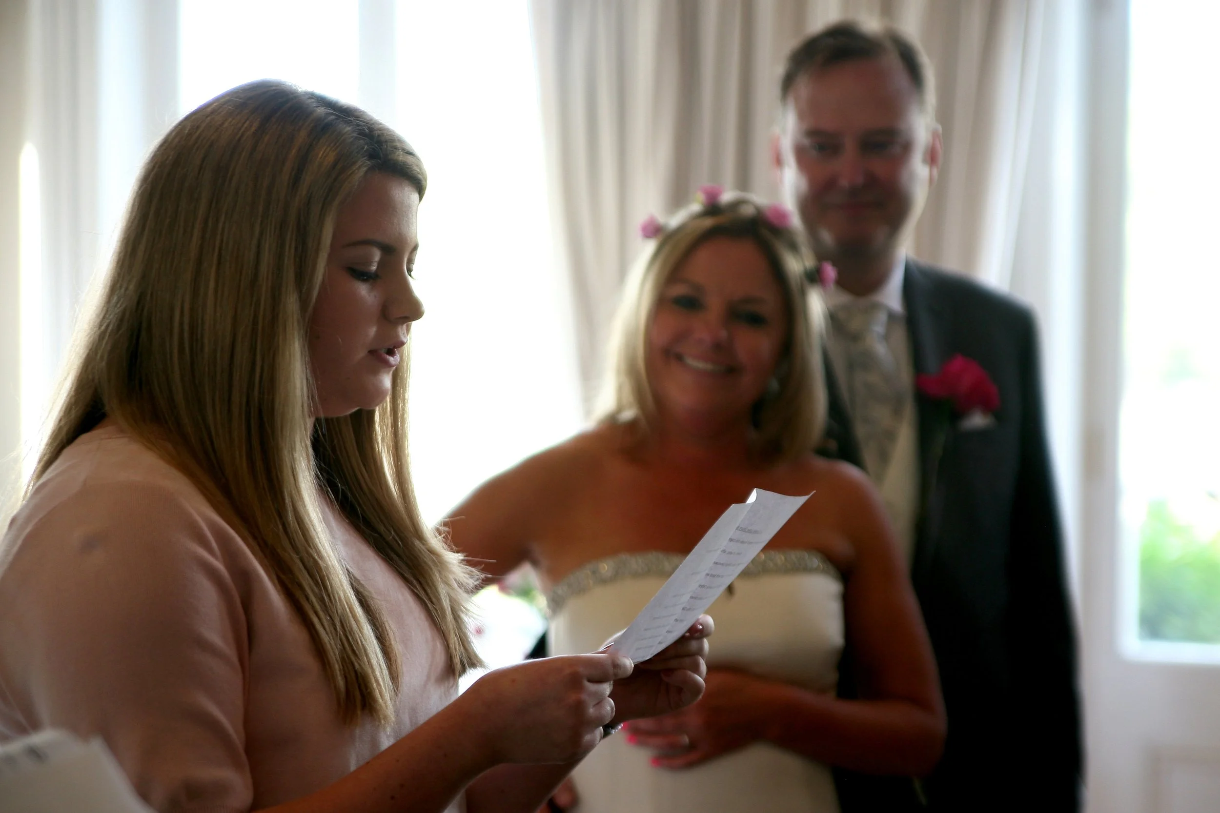 A young woman reading a speech during a wedding ceremony, with a smiling bride and groom standing behind her in a room with white curtains.