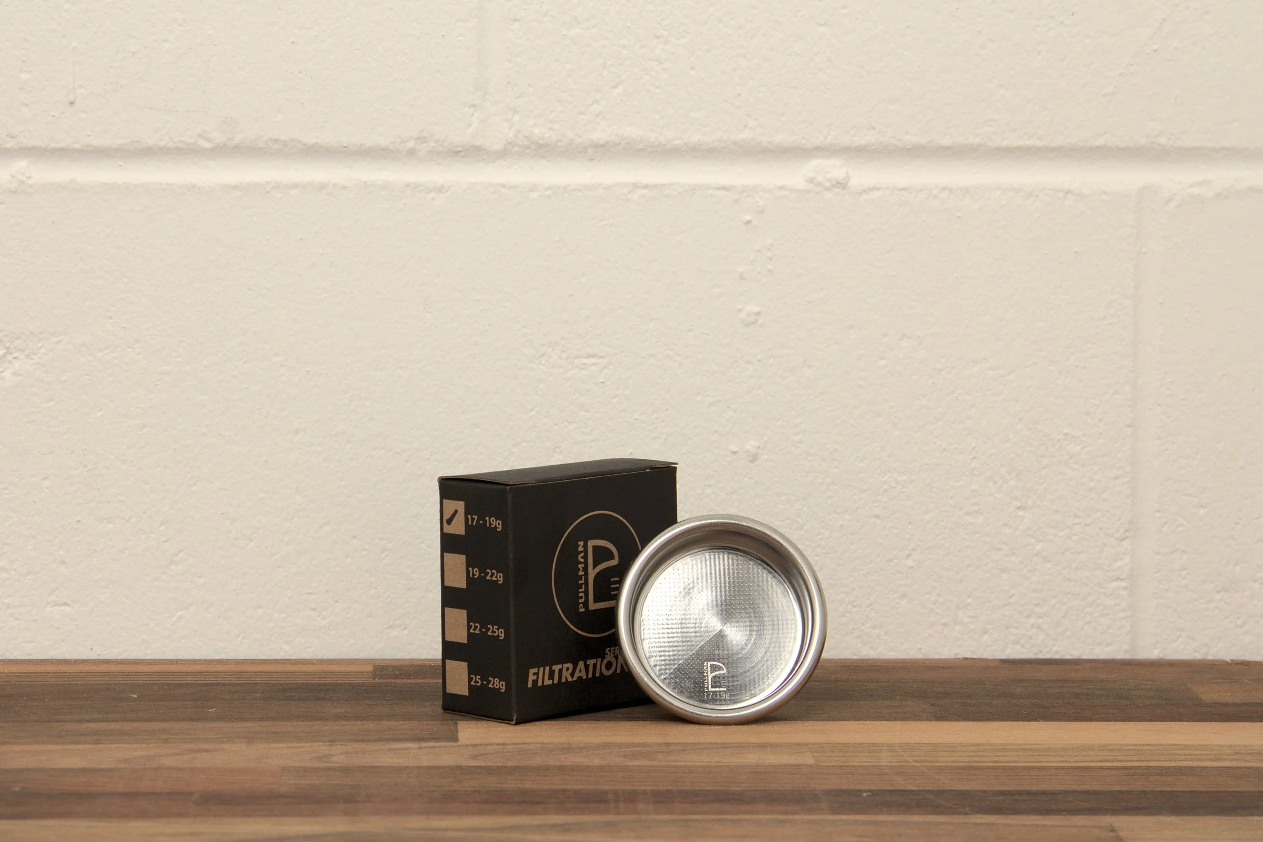 A silver portafilter basket and black box on a wooden surface against a white brick wall background.