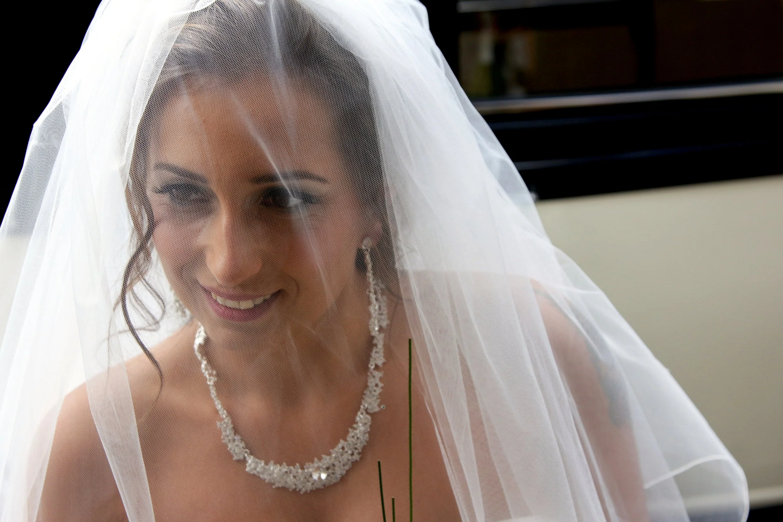 A bride smiling, wearing a white veil, a beaded necklace, and earrings, with dark hair styled in loose waves.