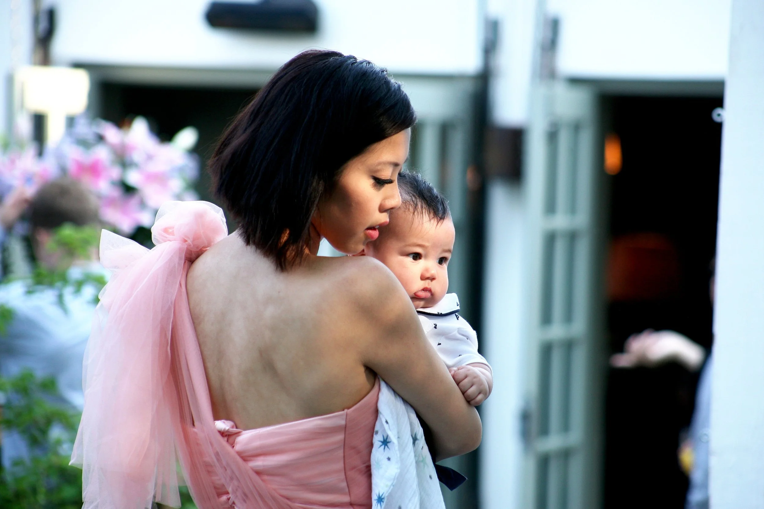 A bridesmaid holding a young child, both looking to the side, with a soft background of flowers and a building.