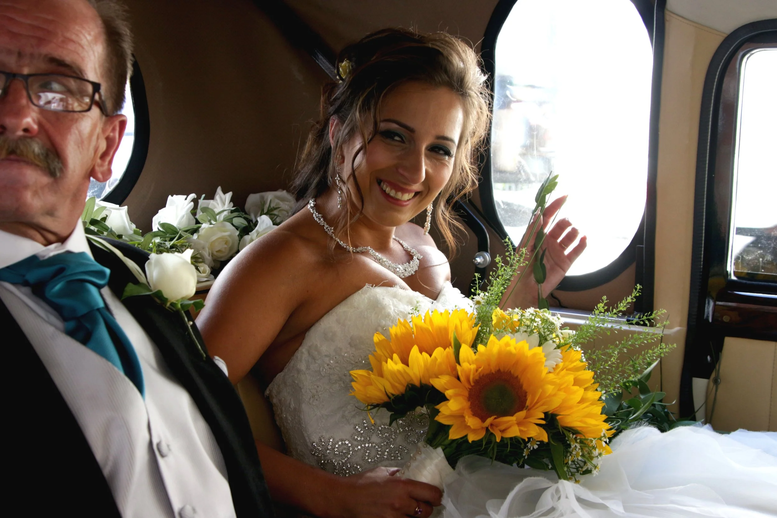 A smiling bride in wedding gown holding a bouquet of sunflowers, seated next to an older man in a tuxedo with a blue bow tie, inside a vintage car.