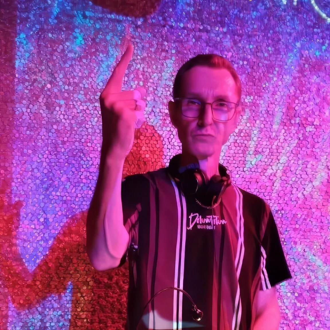 Dean Charles with glasses and headphones around neck, pointing upward with index finger, standing against a colorful, textured wall illuminated in pink and purple lights.