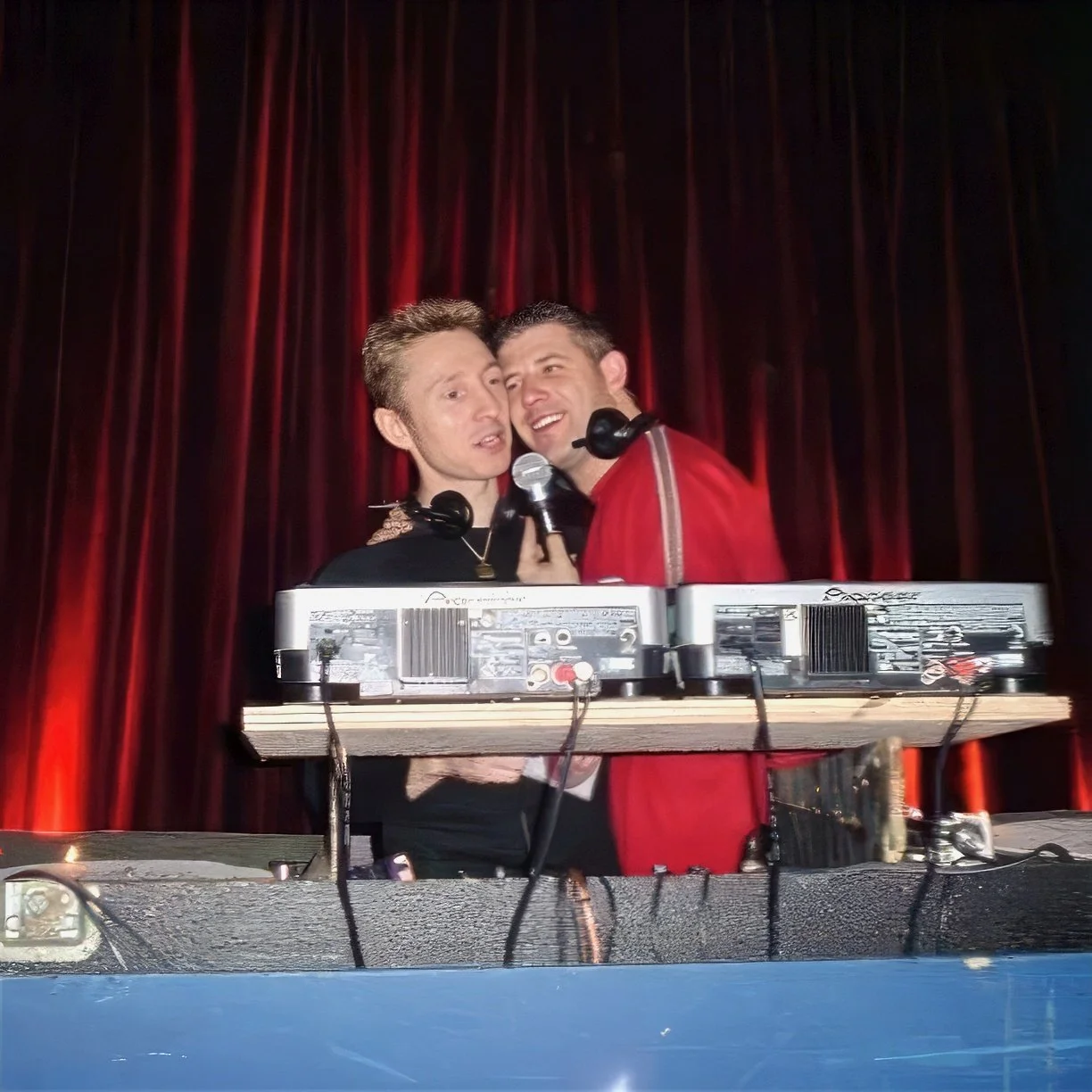 Dj Dean Charles performing on stage with a keyboard and microphones in front of red curtains, sharing a close moment.