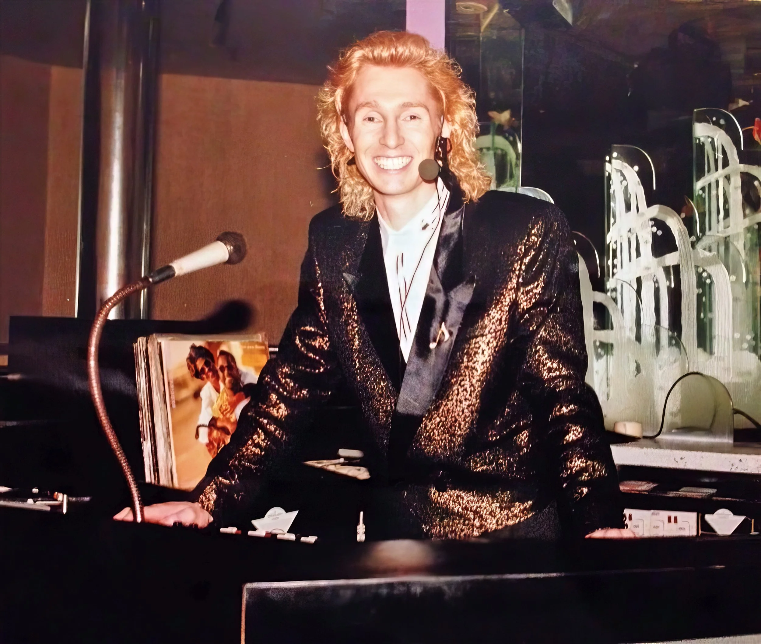 DJ with curly blonde hair smiling and standing in a DJ booth, wearing a black and gold shiny jacket, with a microphone headset, in a music or entertainment venue.