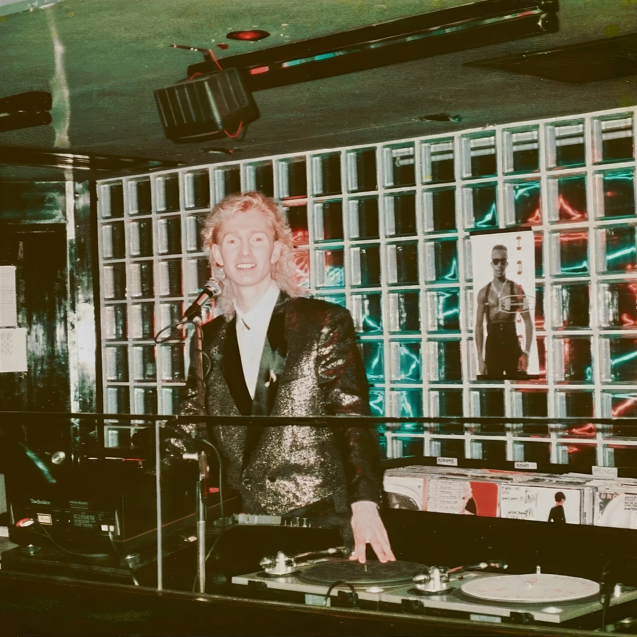 A male DJ with curly blonde hair in a black jacket stands behind turntables with a microphone in front. The background features a wall of square glass blocks and neon lights, with a poster of a MC Hammer wearing sunglasses and a tank top.