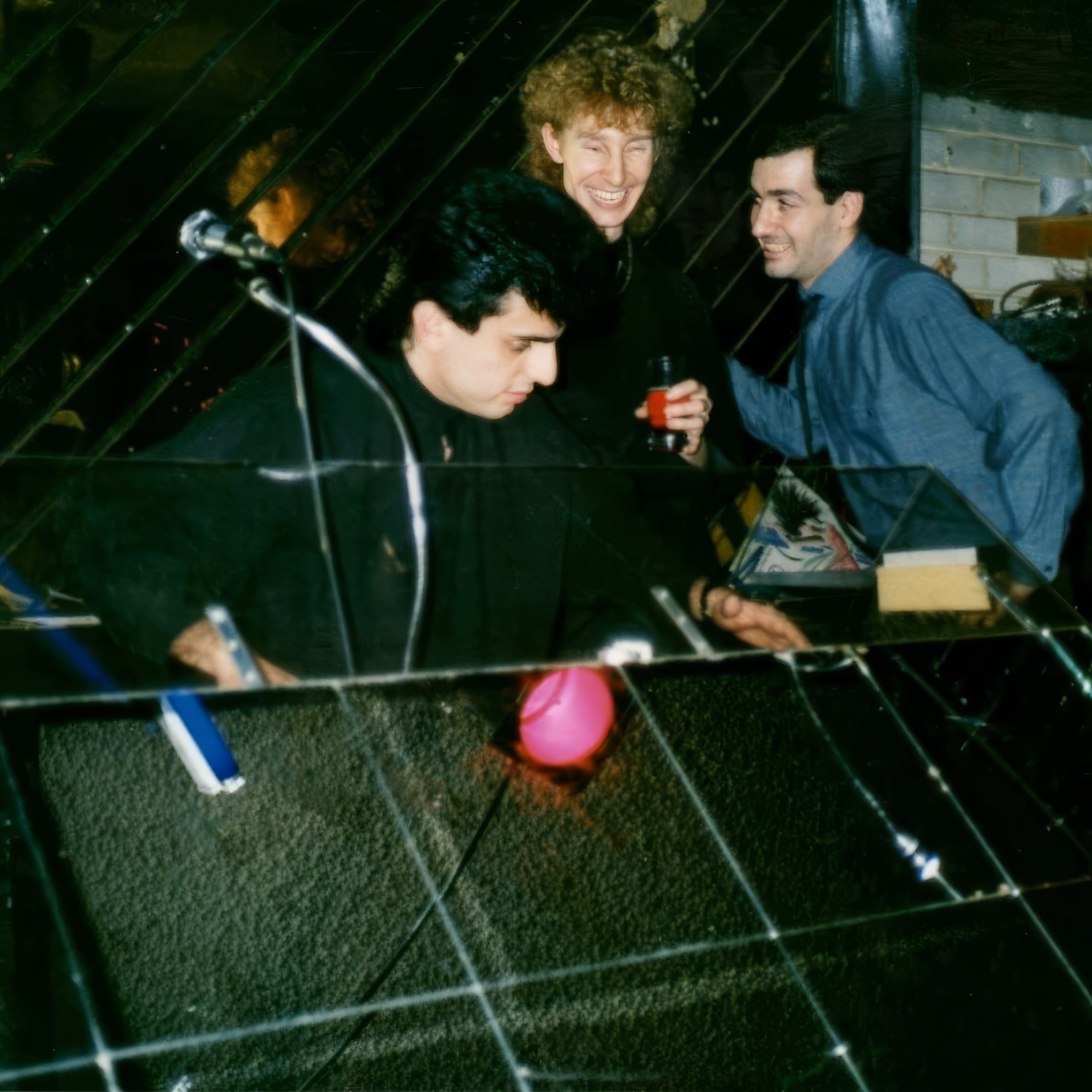 Three men at a party or gathering; one with curly hair smiling and holding a drink, one with dark hair looking down, and another with black hair leaning toward the others. The image is taken through a glass or plastic reflective surface with a pink o