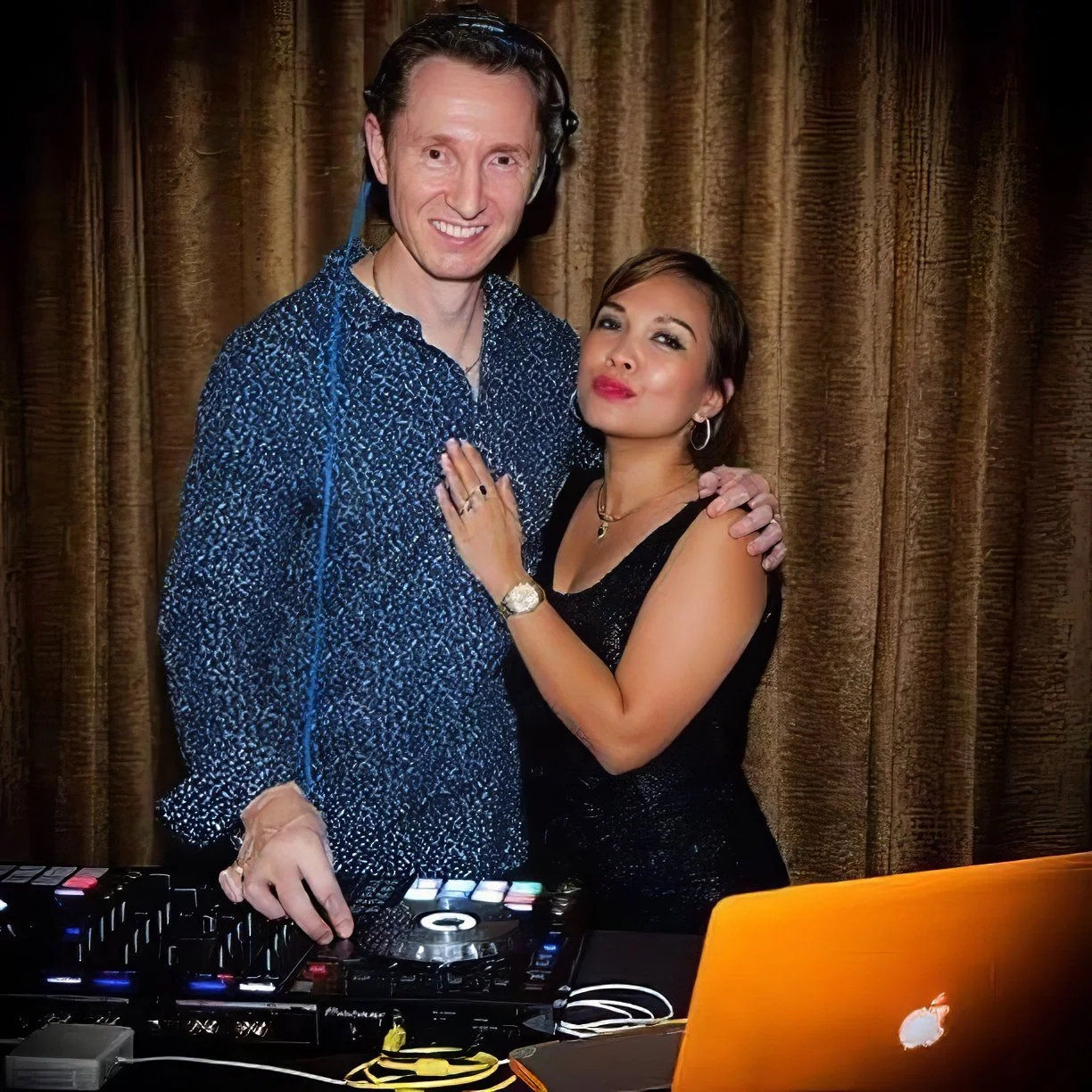 Dean Charles and his wife pose together at an event, with DJ equipment and a laptop in front of them and a brown curtain backdrop behind. Dean is wearing headphones and a blue patterned shirt and his wife is in a black dress with jewelery.