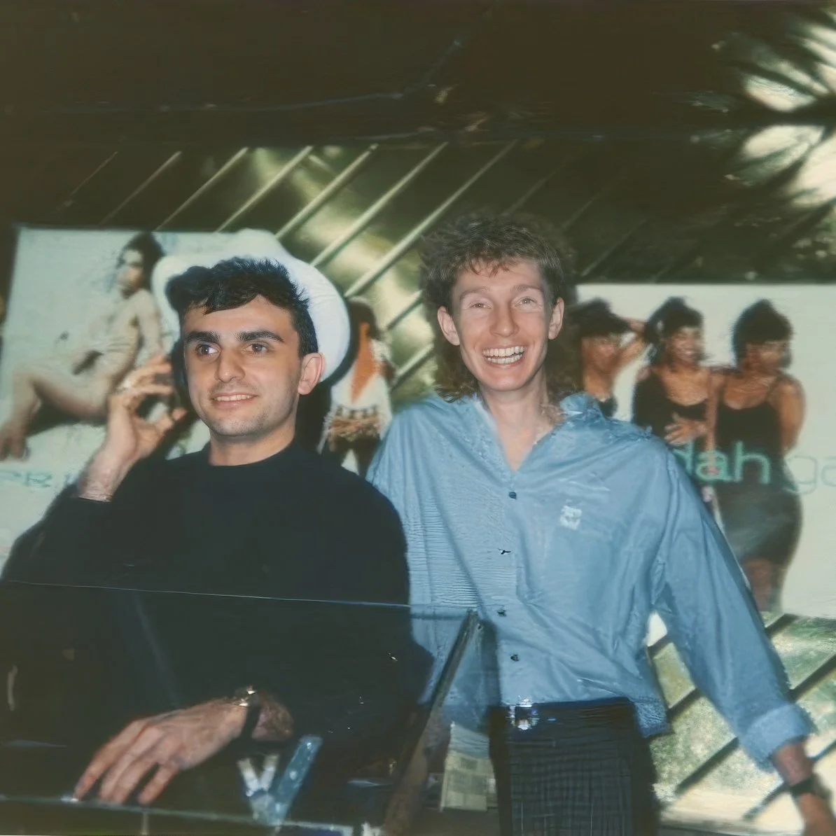 Two men are standing in front of a large colorful poster at an event, one is talking on a mobile phone and the other is smiling widely for the camera. The poster behind them features people in swimsuits on a beach.