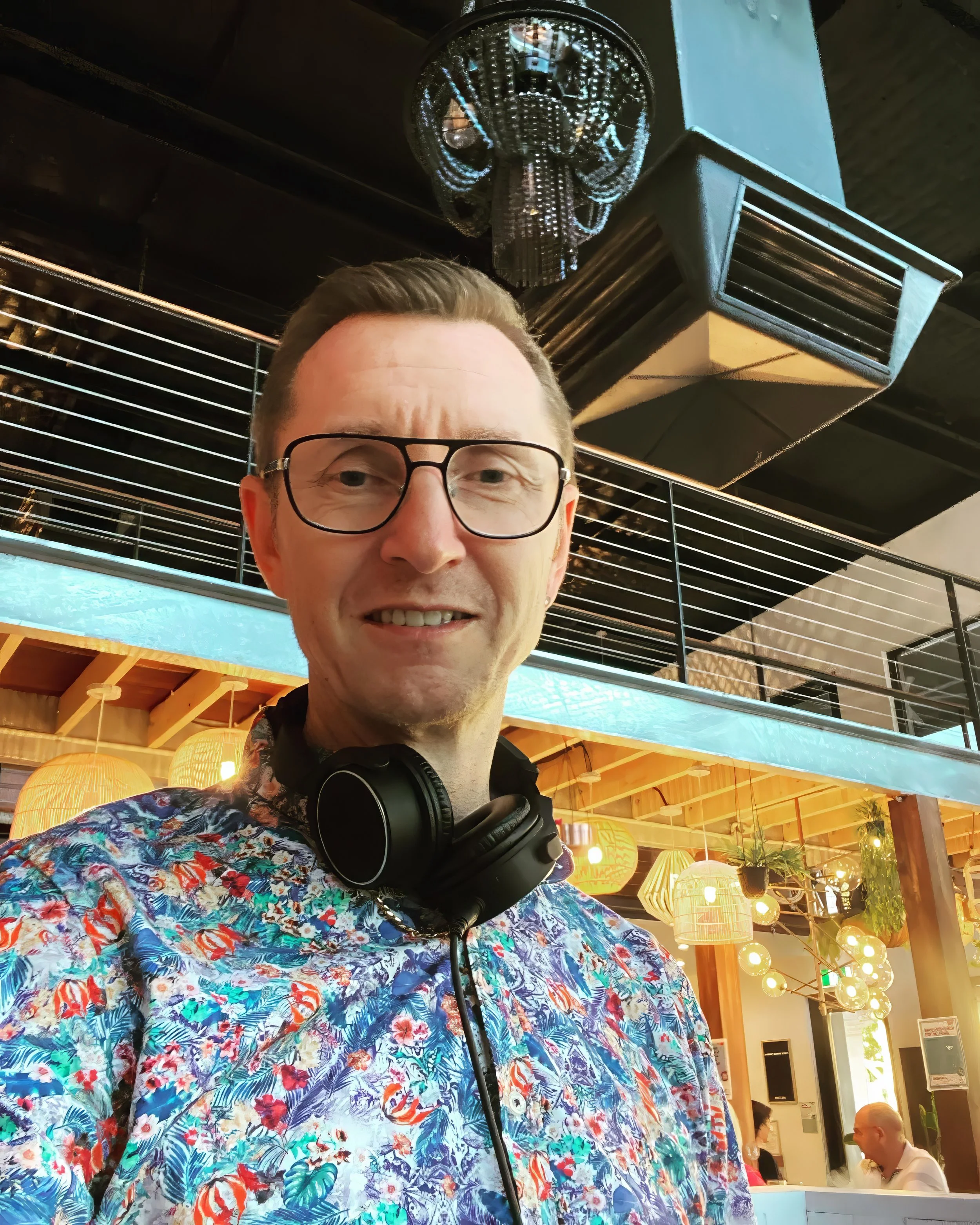 Dean Charles wearing glasses and a colorful floral shirt with headphones around his neck taking a selfie inside Soul bar, decorated restaurant or cafe with hanging lights, plants and other patrons in the background.