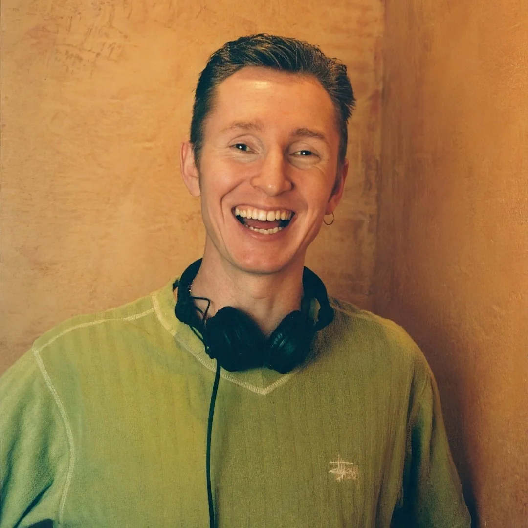 DJ Dean Charles in 2000 at The Church club, wearing a green sweater and black headphones around his neck, smiling broadly in front of a textured beige wall.